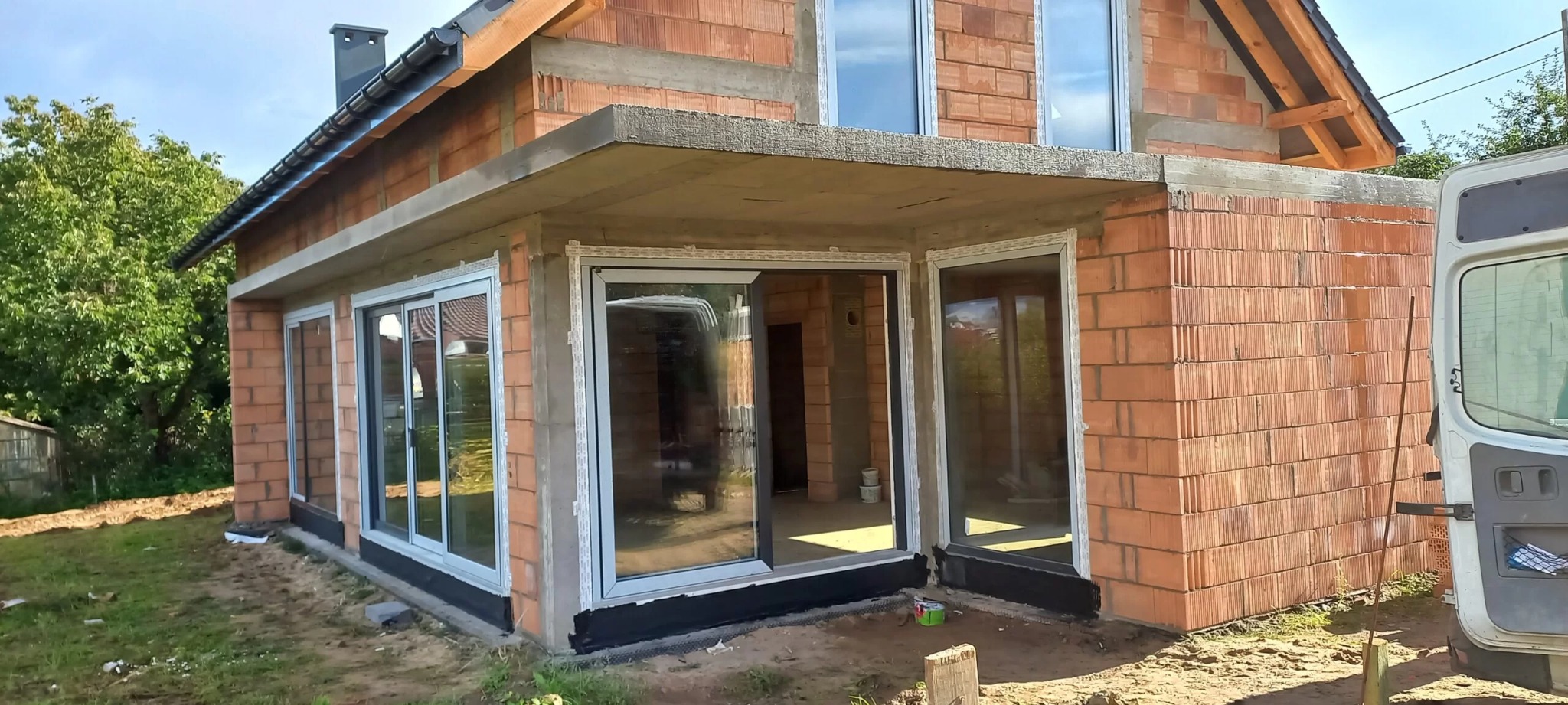 Nowoczesna bryła domu w trakcie budowy z zamontowanymi przesuwnymi oknami aluminiowymi i przygotowanymi otworami na kolejne okna, widoczne materiały budowlane i fragment samochodu dostawczego.