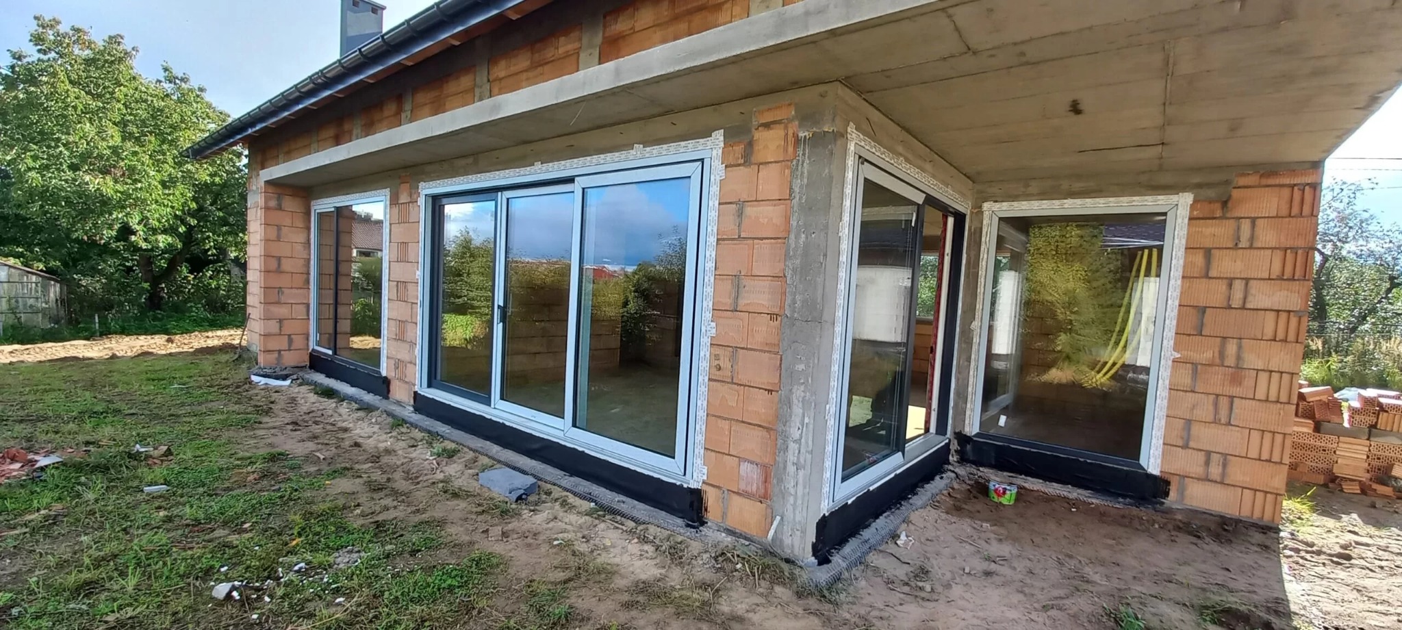 Dom w stanie surowym otwartym z zamontowanymi dużymi przesuwnymi oknami z szarymi ramami, widoczne cegły i betonowa konstrukcja dachu.