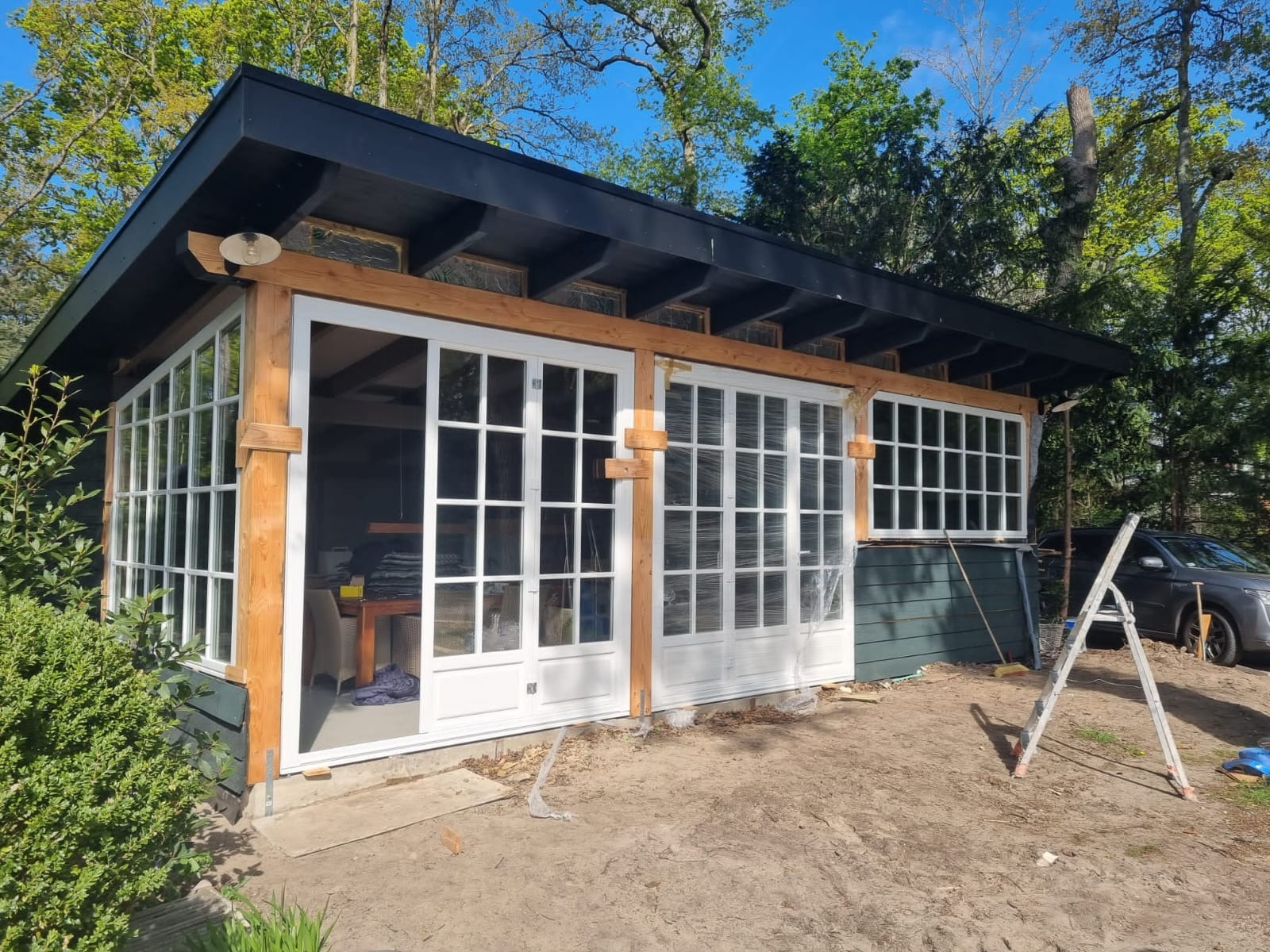 Nowoczesna, przeszklona weranda w trakcie budowy z białymi ramami okiennymi i drzwiowymi, zabezpieczonymi folią, widoczna drabina i samochód w tle.