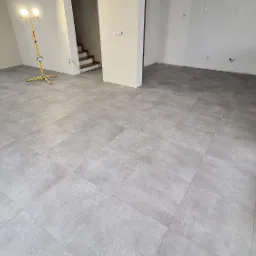 Wyłożona szarą, matową płytką podłoga w jasnym, przestronnym pokoju z widocznymi schodami w tle i lampą budowlaną.