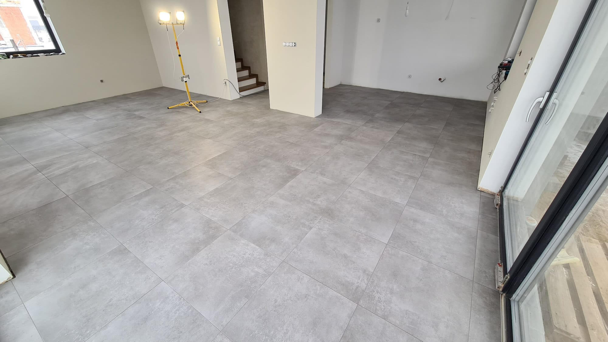 Wyłożona szarą, matową płytką podłoga w jasnym, przestronnym pokoju z widocznymi schodami w tle i lampą budowlaną.