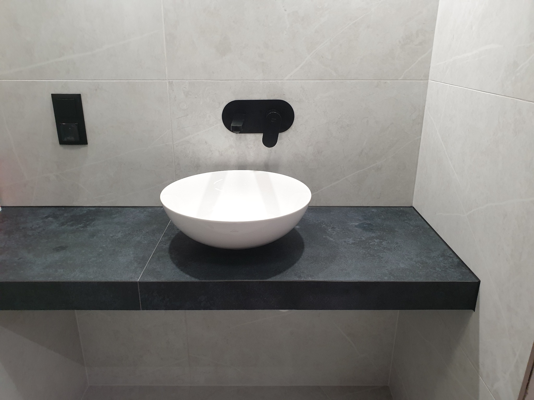 Nowoczesna łazienka z białą, okrągłą umywalką nablatową, czarną baterią ścienną i czarnym blatem z kamienia, na tle płytek imitujących marmur.