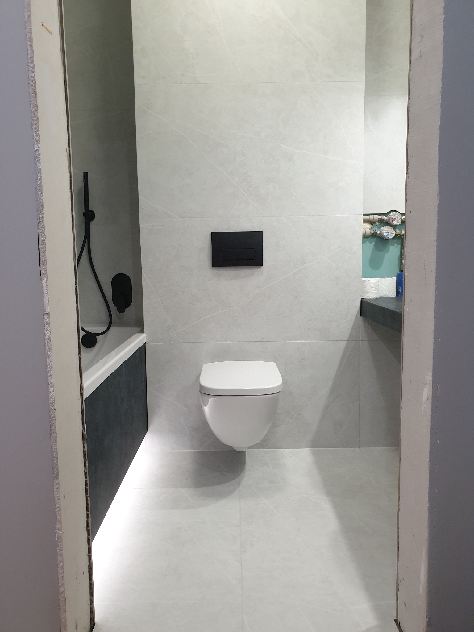 Nowoczesna łazienka z białymi płytkami imitującymi marmur, wisząca toaleta z czarnym przyciskiem spłukującym, czarna armatura prysznicowa i fragment wanny z ciemnoszarą obudową, widok zza futryny...