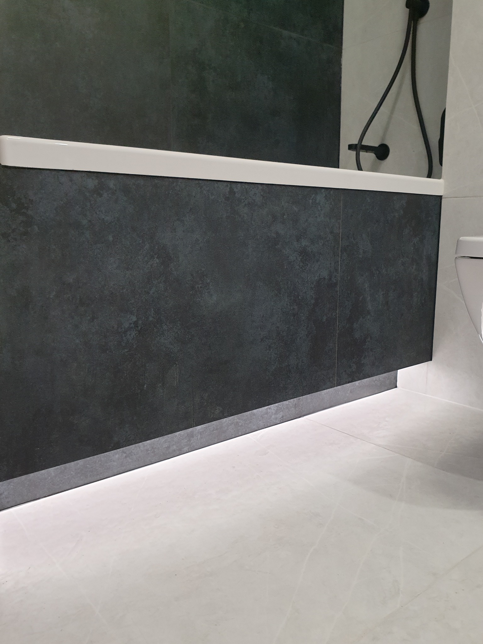 Nowoczesna łazienka z ciemnymi, strukturalnymi płytkami na wannie, podświetleniem LED u dołu i jasnymi marmurkowymi płytkami podłogowymi, ukazująca minimalistyczny design.