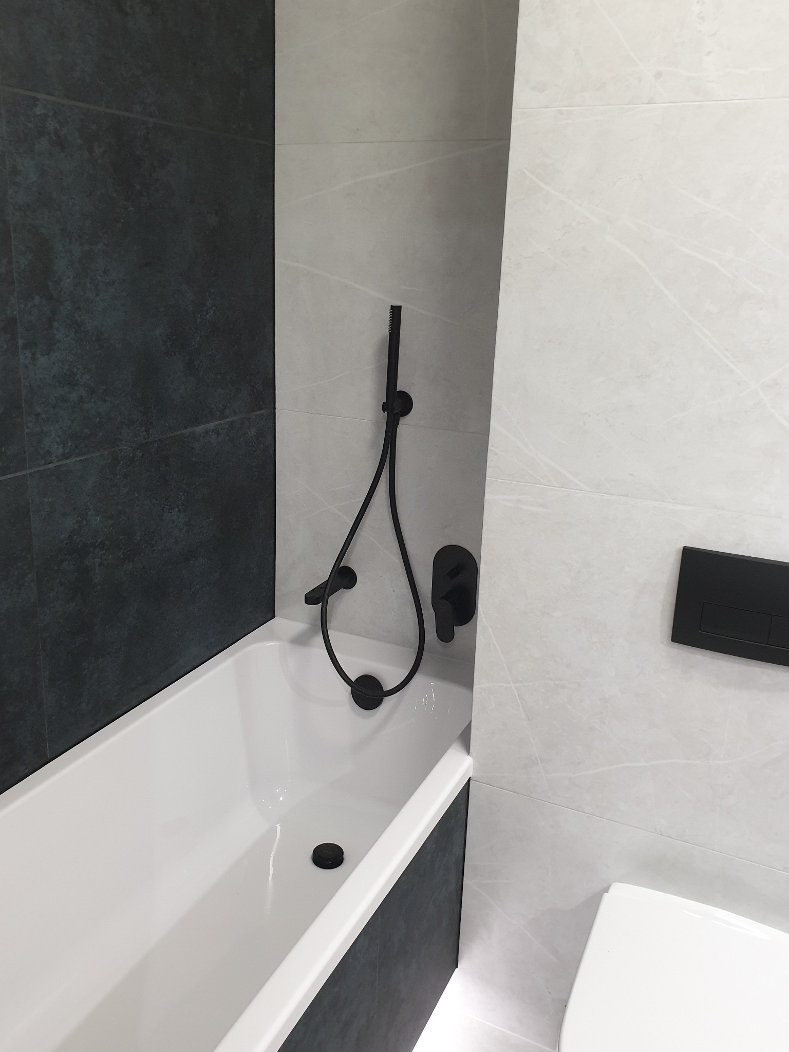 Nowoczesna łazienka z białą wanną, czarną armaturą i szaro-białymi płytkami imitującymi marmur.