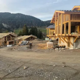 Wczesny etap budowy kilku domów z drewnianą konstrukcją szkieletową, widok na wzgórza w tle, teren budowy z gruzem i śladami opon.