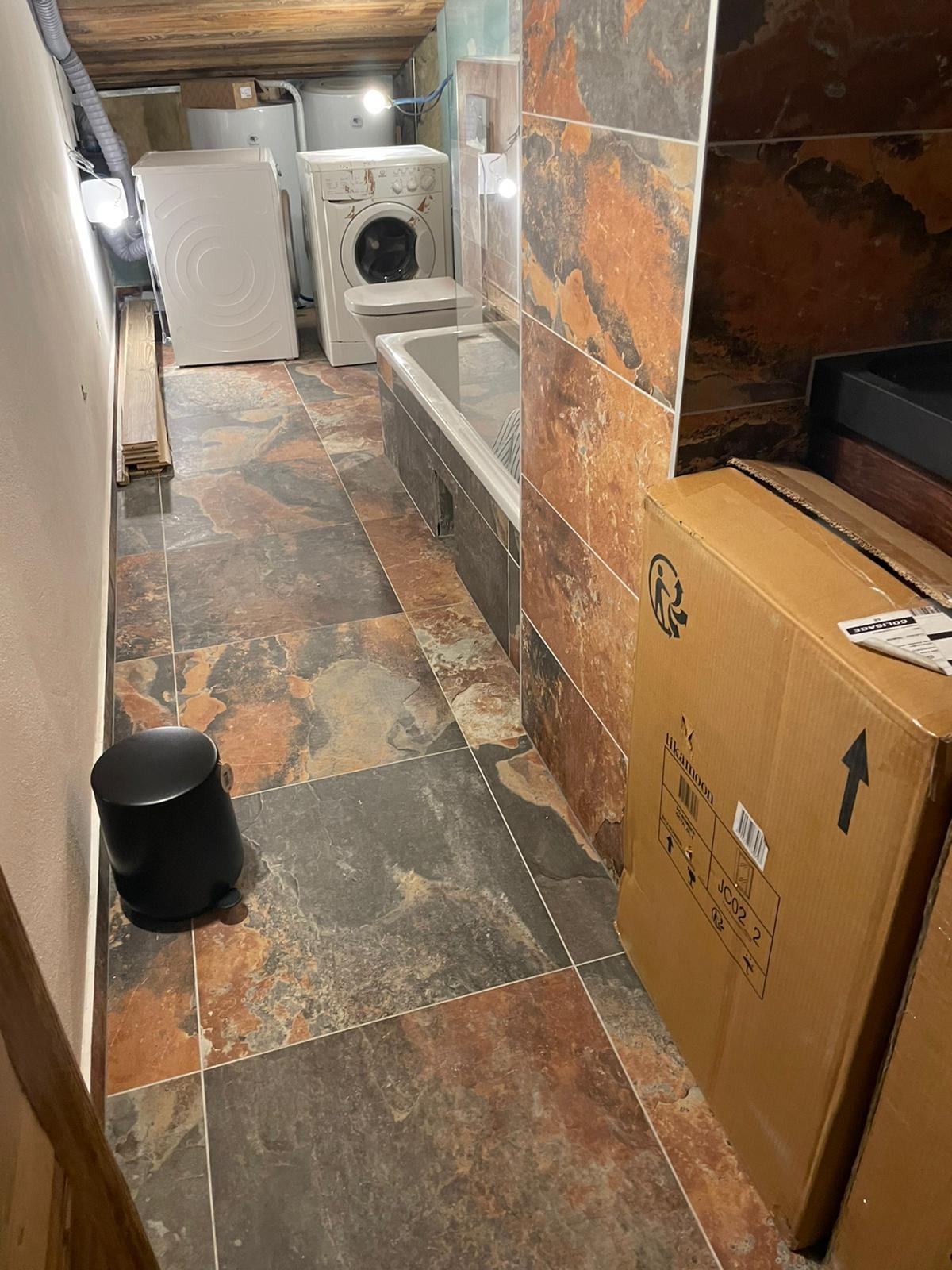 Wnętrze łazienki z płytkami imitującymi kamień, widoczne pralka, sedes, wanna, kosz na śmieci i kartonowe pudła.