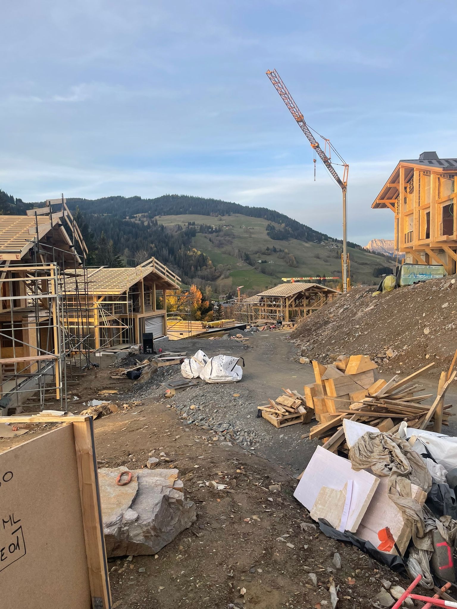 Budowa kilku domów szkieletowych z drewna na tle górskim, widoczny żuraw, materiały budowlane i częściowo wykończone konstrukcje.