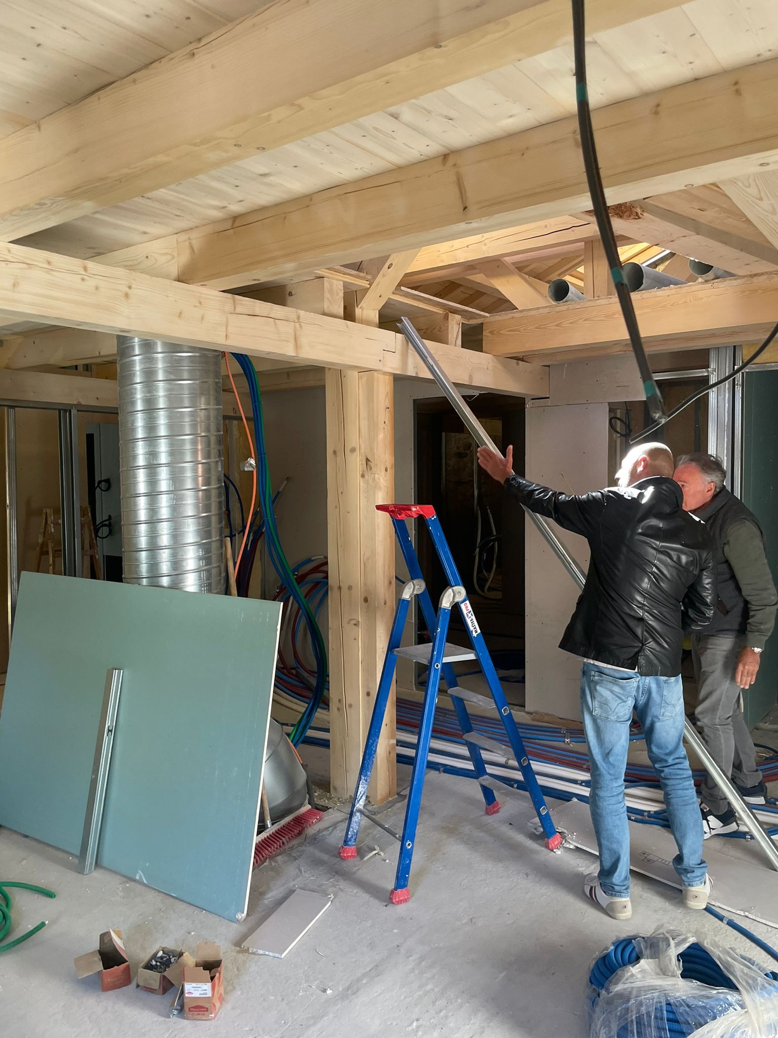 Wnętrze w trakcie budowy z drewnianą konstrukcją stropu, widoczne instalacje, płyty gipsowe i dwaj mężczyźni omawiający montaż metalowego profilu na tle niebieskiej drabiny.
