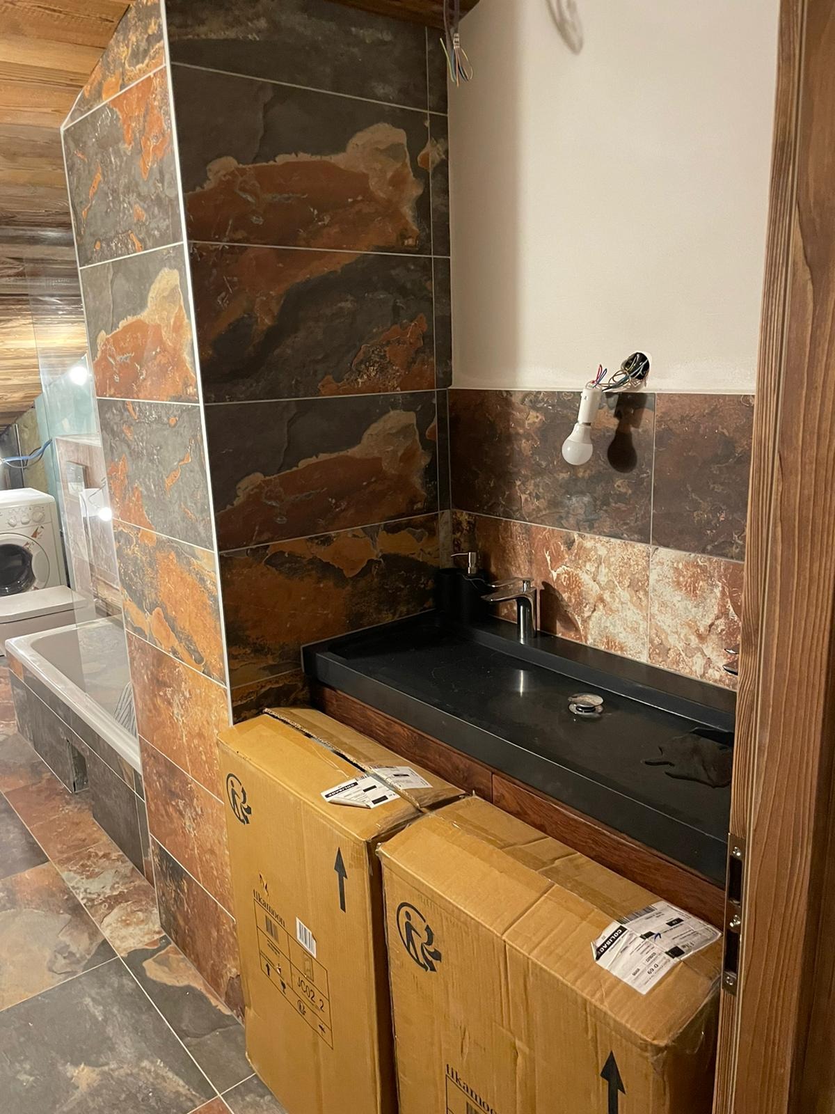 Łazienka w trakcie remontu: czarna umywalka z baterią, okładzina ścienna z płytek imitujących kamień, niezabezpieczone przewody oświetleniowe wystające ze ściany, kartony z wyposażeniem na podłodze.
