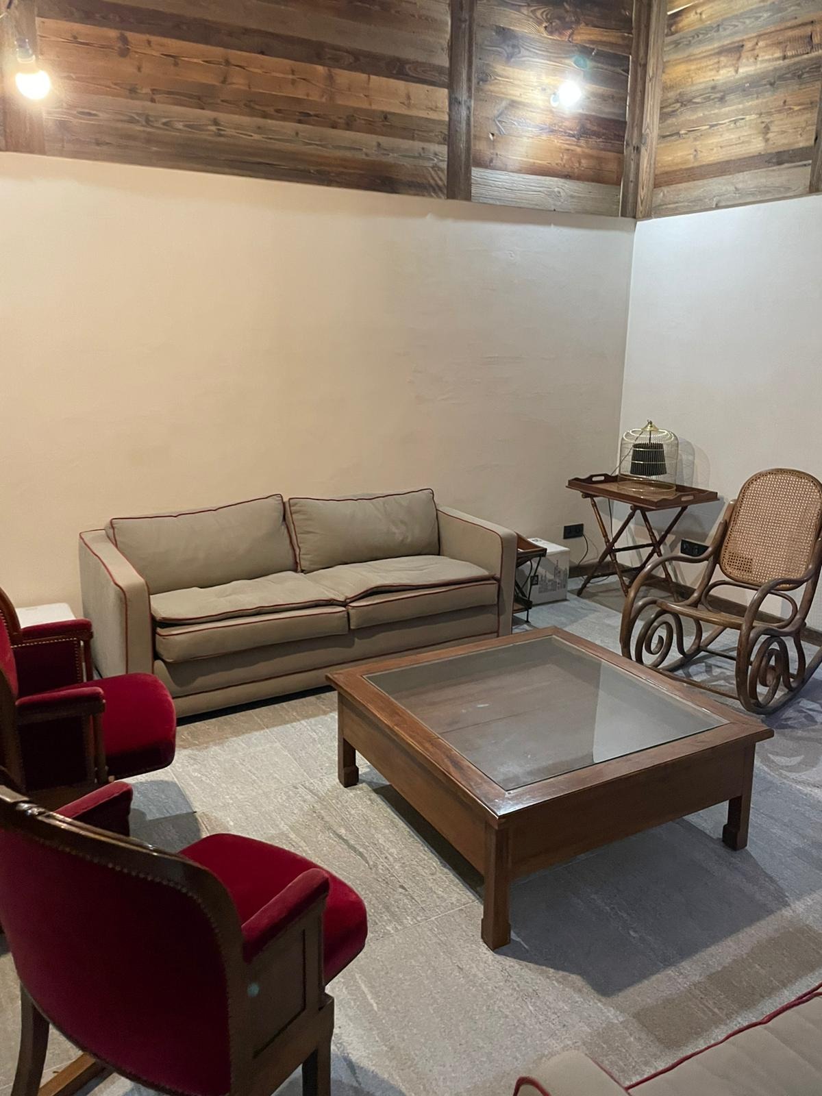 Wnętrze salonu w domu z bali z drewnianym sufitem, beżową sofą z czerwonymi lamówkami, dwoma czerwonymi fotelami, drewnianym stolikiem kawowym ze szklanym blatem i wiklinowym fotelem bujanym.