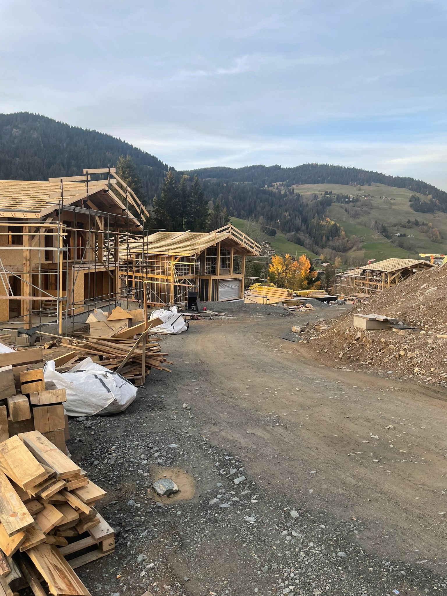 Budowa szkieletowych domów w górskim krajobrazie, widoczne drewniane konstrukcje, rusztowania, materiały budowlane i pagórkowate tło.