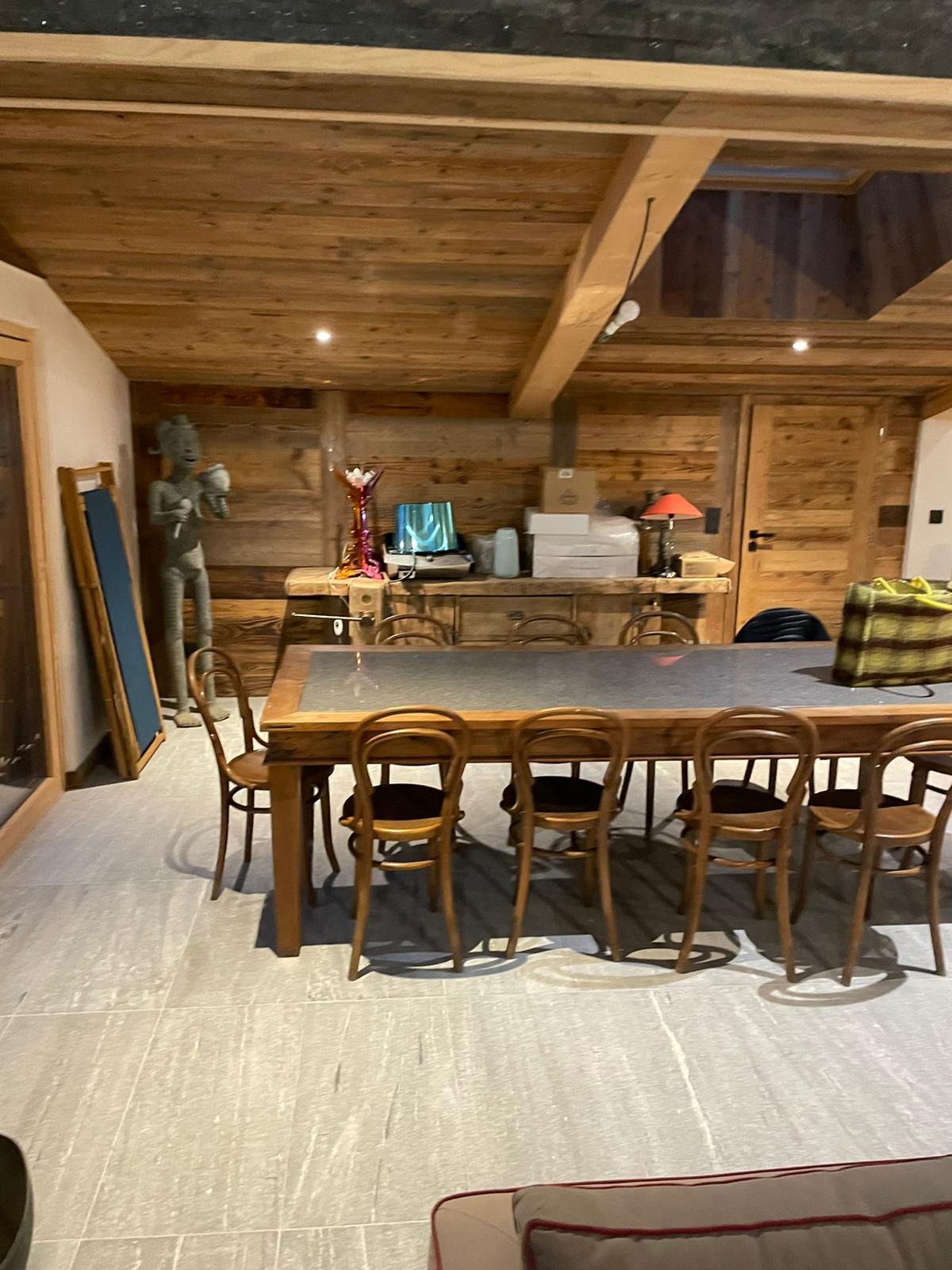 Wnętrze jadalni w domu z drewnianymi ścianami i sufitem, duży stół z krzesłami, rzeźba postaci stojąca obok tablicy, oświetlenie punktowe i okno dachowe.