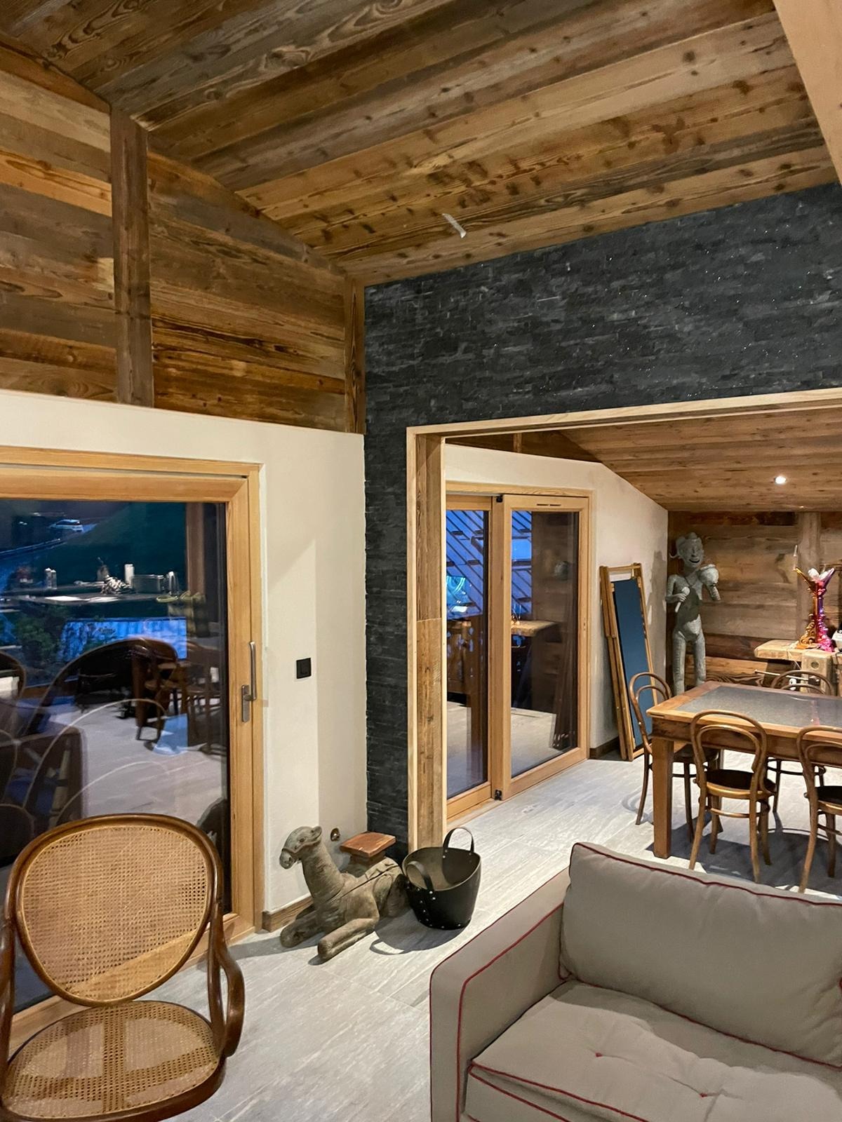 Wnętrze domu z drewnianym wykończeniem ścian i sufitu, widoczne okna z widokiem na zewnątrz, drewniany stół z krzesłami oraz dekoracyjne rzeźby.