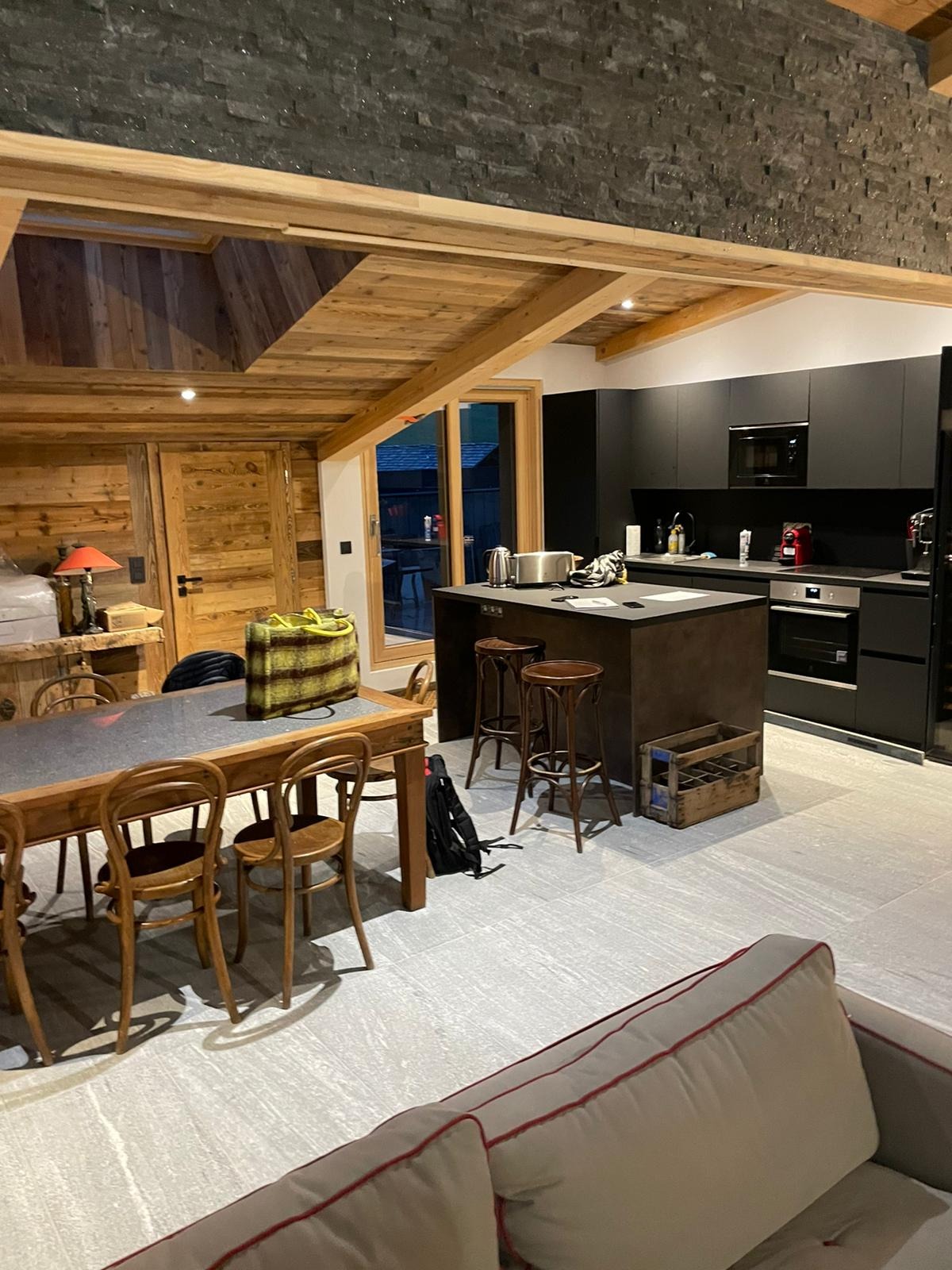 Wnętrze domu z bali z drewnianym stropem, widoczna kuchnia z czarnymi szafkami, wyspa kuchenna z hokerami i duży stół z krzesłami.