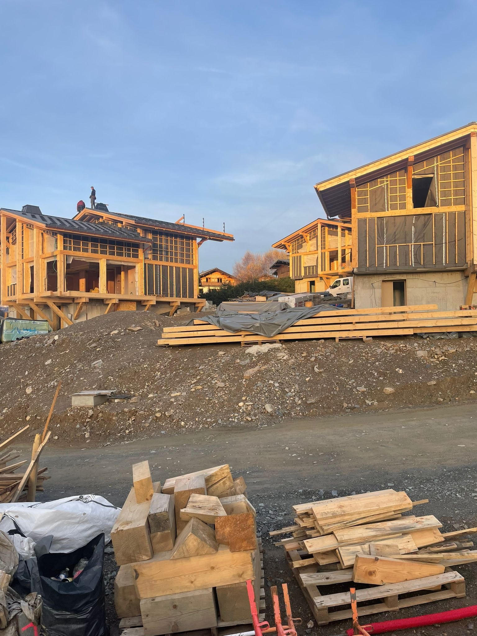 Szkieletowa konstrukcja dwóch budynków w trakcie budowy, drewniane belki, widoczni pracownicy na dachu jednego z budynków, sterta ziemi i materiałów budowlanych na pierwszym planie.