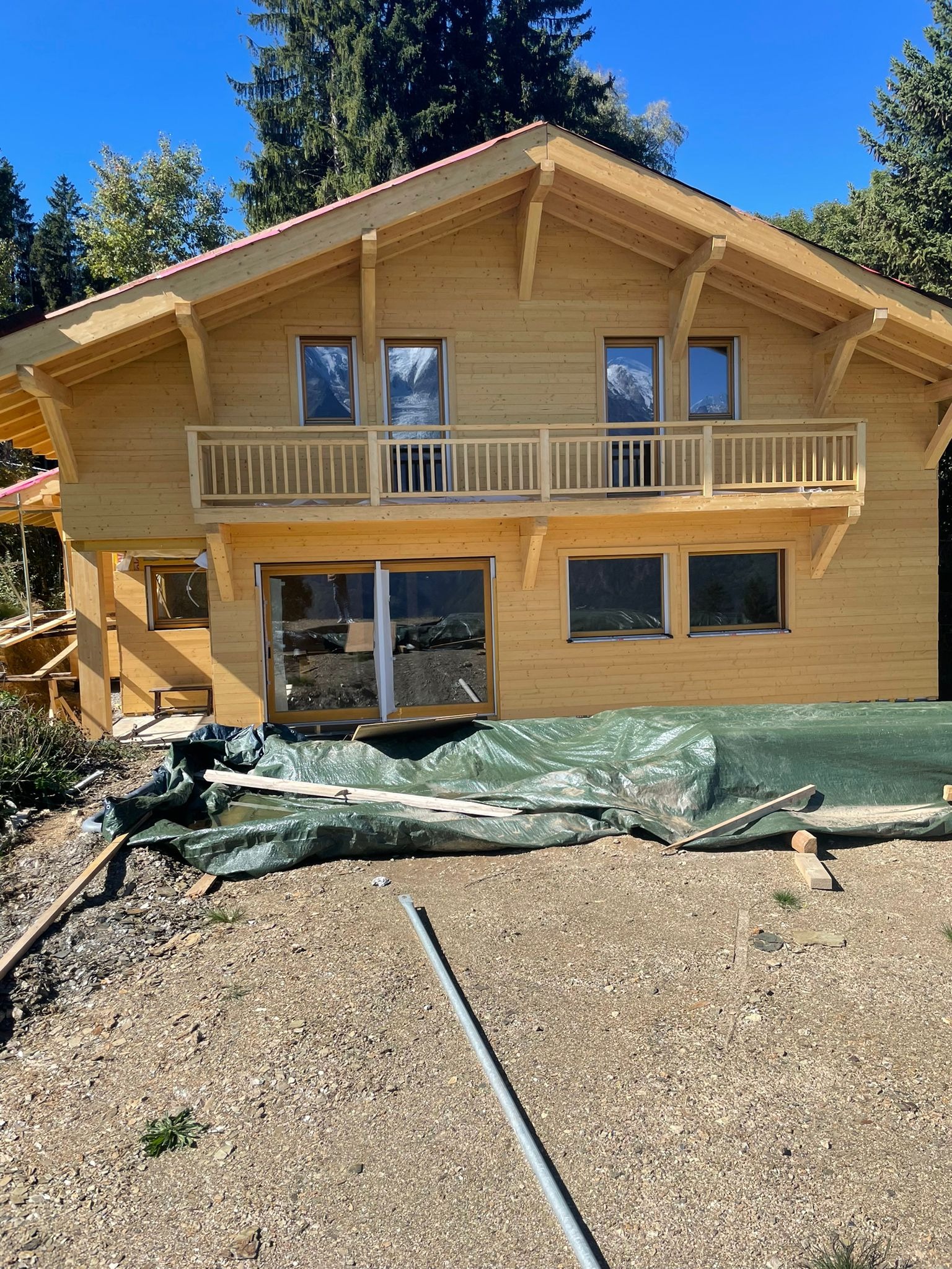 Surowy, dwukondygnacyjny dom z drewna w trakcie budowy, z balkonem na piętrze, zabezpieczony zieloną plandeką na ziemi, w tle widoczne drzewa.