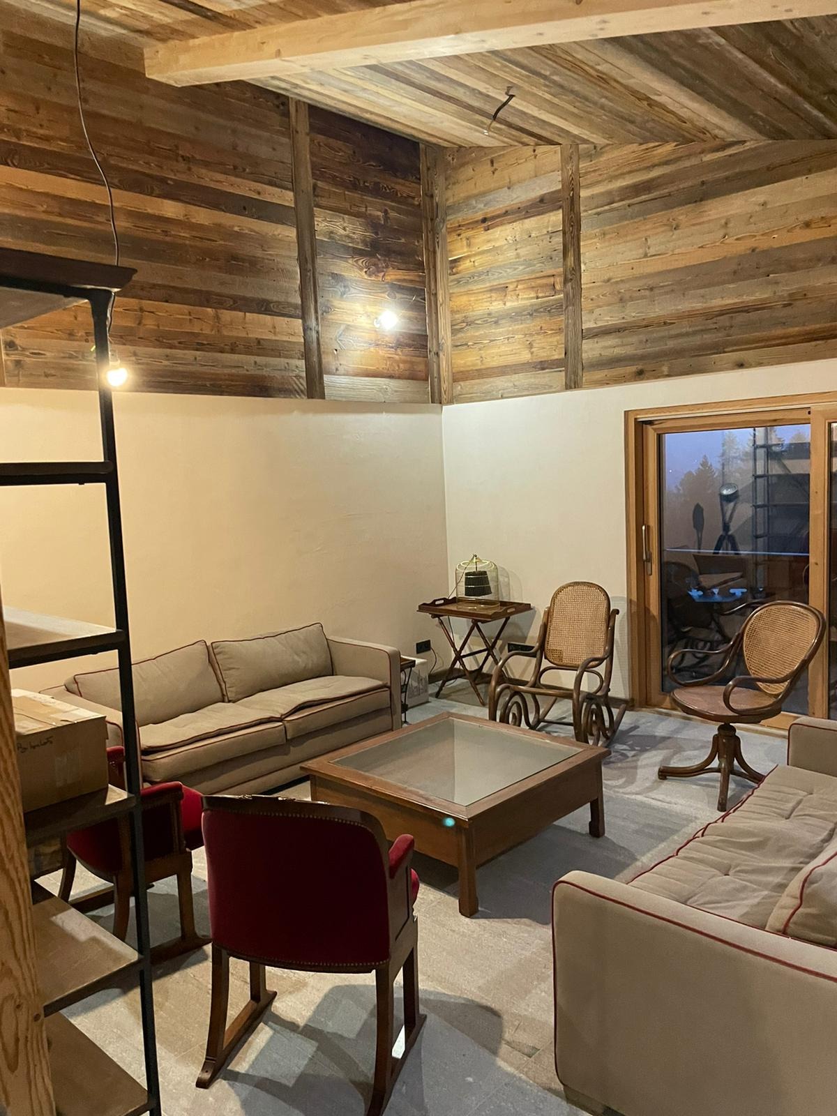 Jasne wnętrze salonu z drewnianym sufitem i ścianami, beżowe sofy z czerwonymi lamówkami, szklany stolik kawowy, wiklinowe krzesła i widok na zewnątrz przez przesuwne drzwi balkonowe.