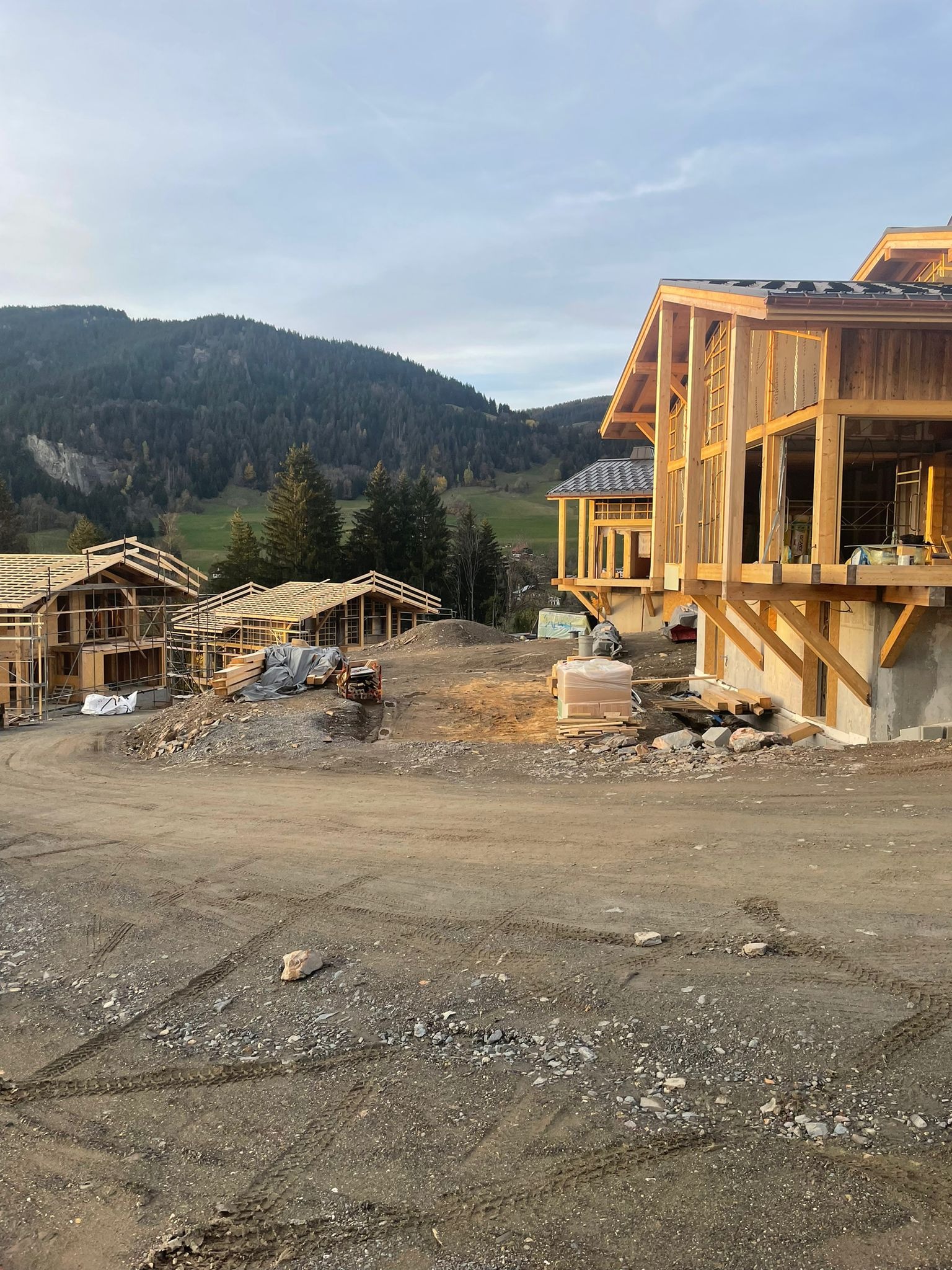 Wczesny etap budowy kilku domów z drewnianą konstrukcją szkieletową, widok na wzgórza w tle, teren budowy z gruzem i śladami opon.