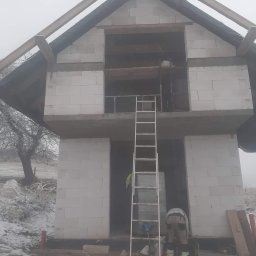 Lukasz - Wznoszenie dwukondygnacyjnego domu z białych bloczków w zimowej scenerii. Widoczna drabina oparta o ścianę, częściowo zamontowane drewniane elementy dachu, oraz pracownik w jasnych spodniach.