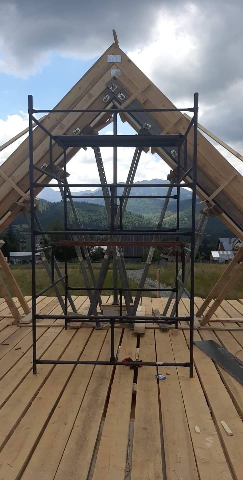 Konstrukcja drewnianego dachu w trakcie budowy, widok na góry przez otwór w dachu, rusztowanie metalowe wewnątrz konstrukcji, deski podłogowe.