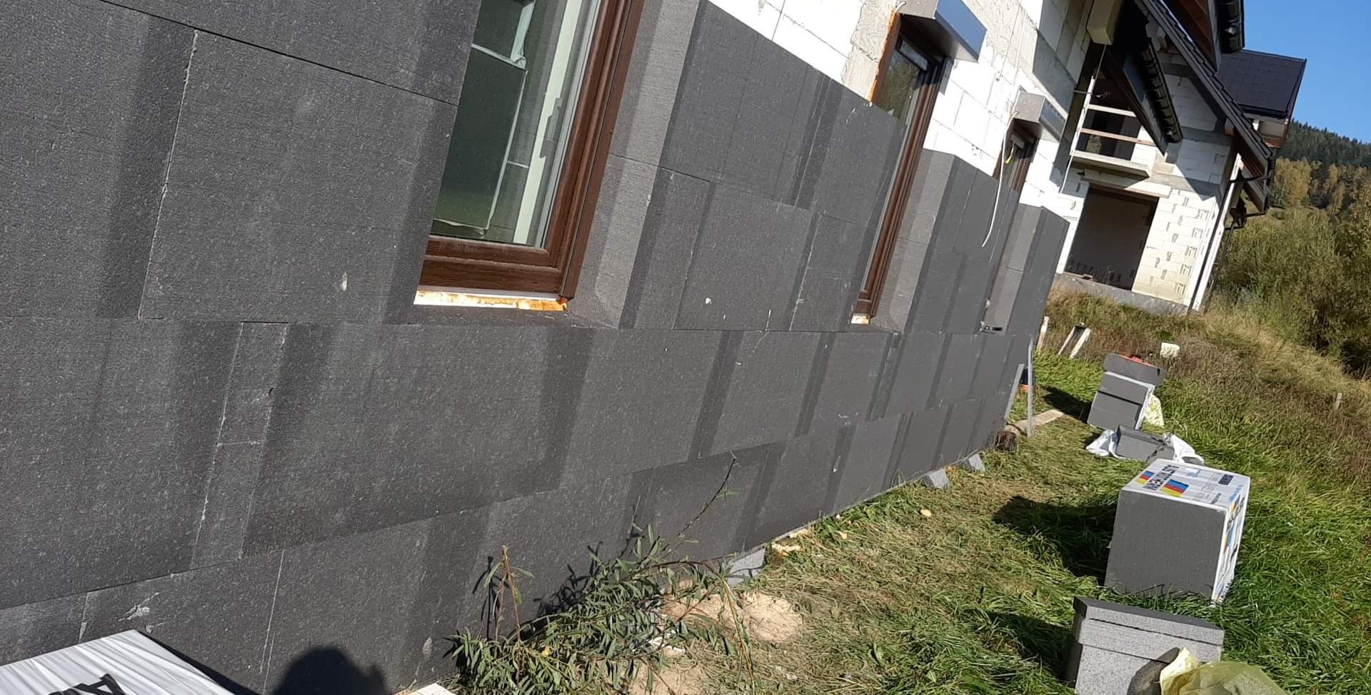 Ocieplanie elewacji domu jednorodzinnego z wykorzystaniem grafitowych płyt styropianowych, widoczne pakiety materiału na trawie oraz fragmenty niezabezpieczonych okien.