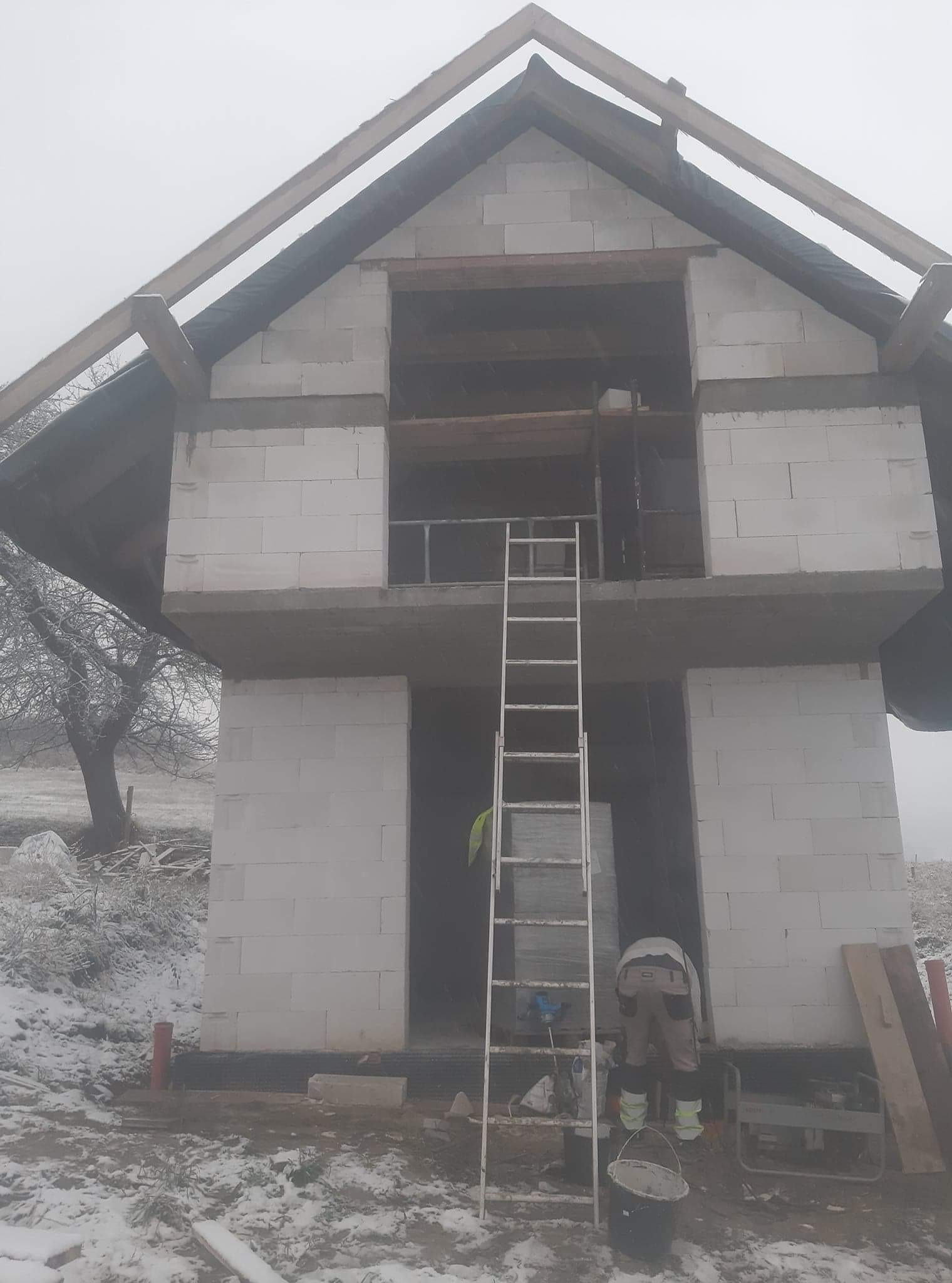 Wznoszenie dwukondygnacyjnego domu z białych bloczków w zimowej scenerii. Widoczna drabina oparta o ścianę, częściowo zamontowane drewniane elementy dachu, oraz pracownik w jasnych spodniach.