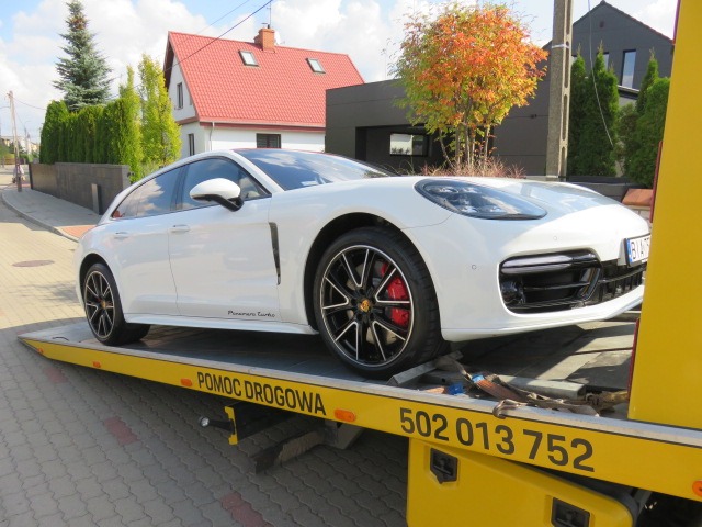 Białe Porsche Panamera Turbo na żółtej platformie pomocy drogowej z widocznym numerem telefonu, na tle ulicy i domów.