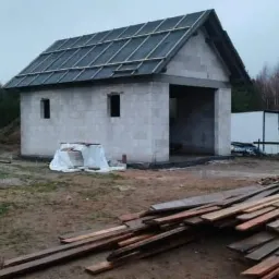 Budowa garażu z pustaków, widoczne drewniane łaty na dachu, teren budowy z hałdą ziemi i stertą desek