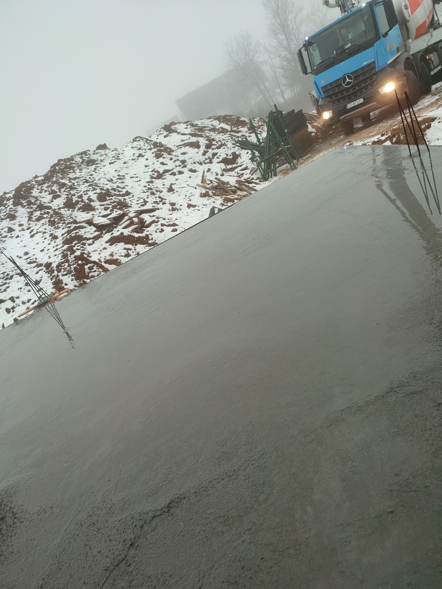 Świeżo wylany, mokry beton na fundamencie domu w pochmurny dzień. W tle widoczna gruszka betoniarki i zasypana śniegiem ziemia.