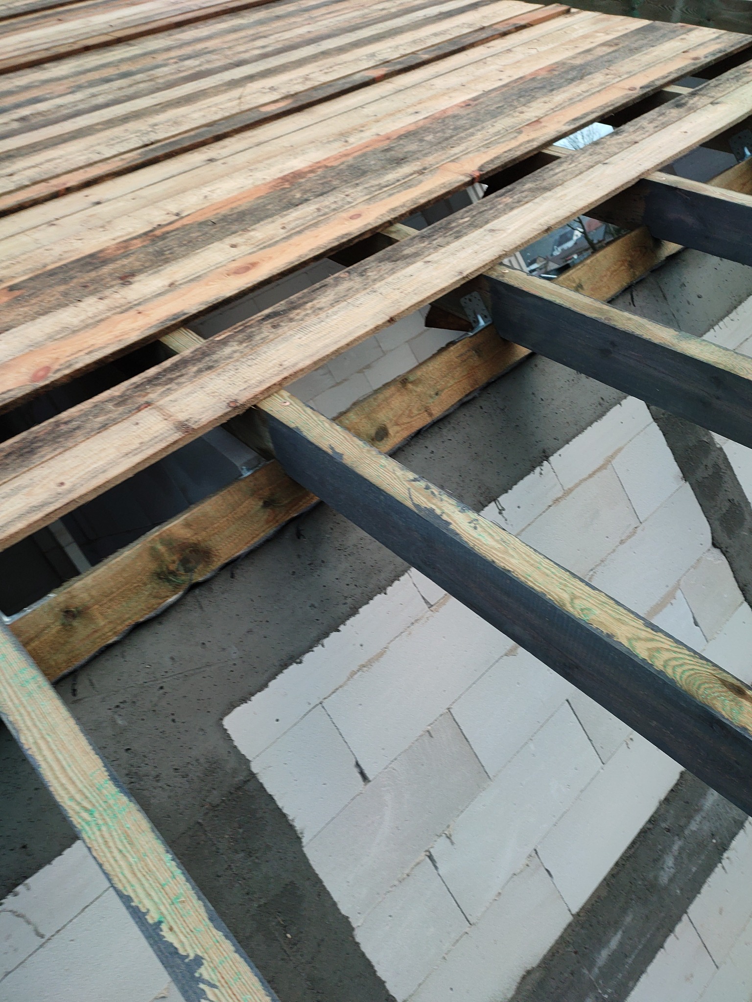 Wznoszenie ścian z bloczków betonowych oraz montaż drewnianej konstrukcji stropu, widoczne elementy mocujące i surowe drewno.