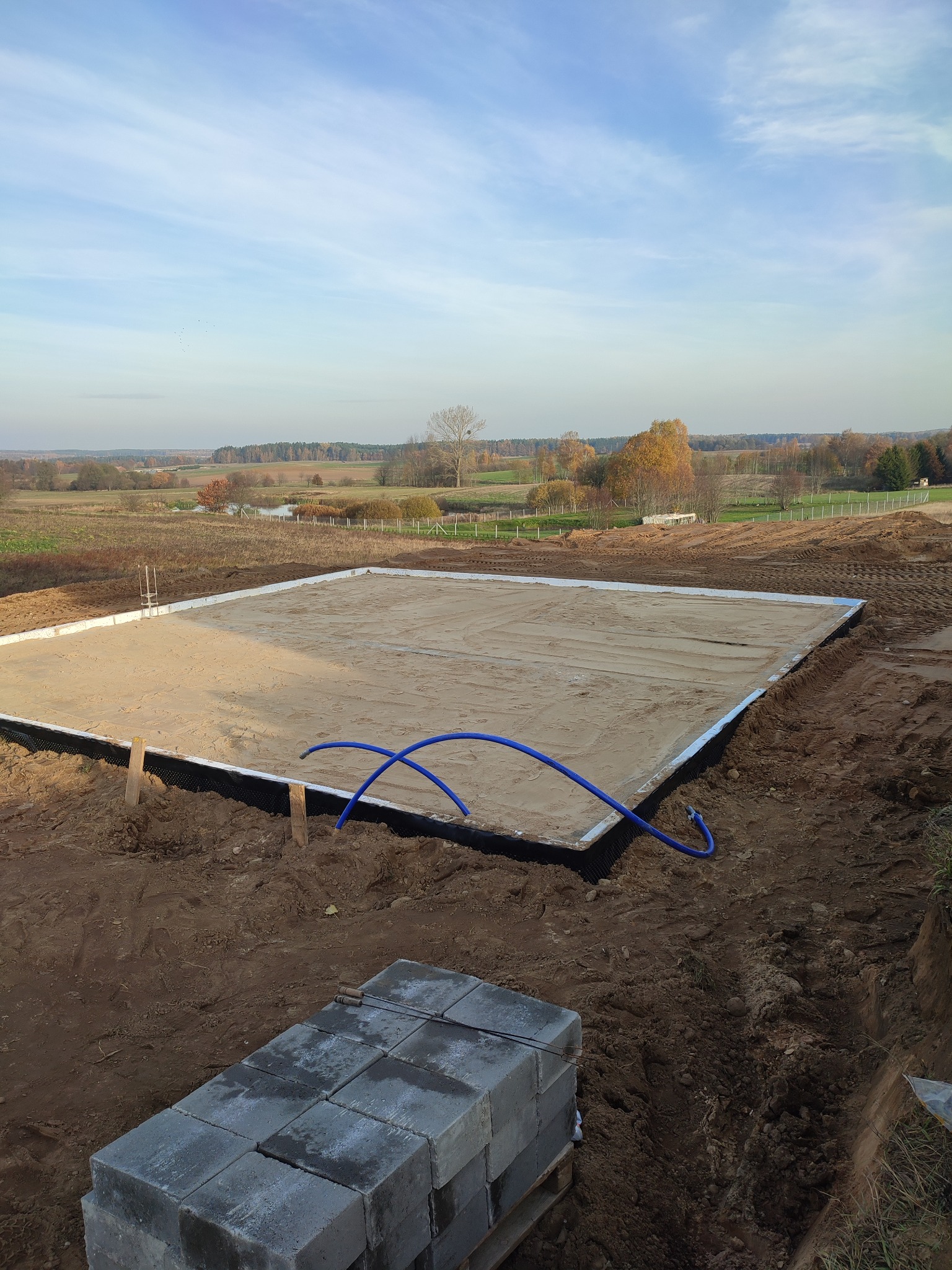 Przygotowany teren pod fundamenty z ułożoną izolacją i zasypany piaskiem, widoczne rury instalacyjne i paleta z bloczkami betonowymi.