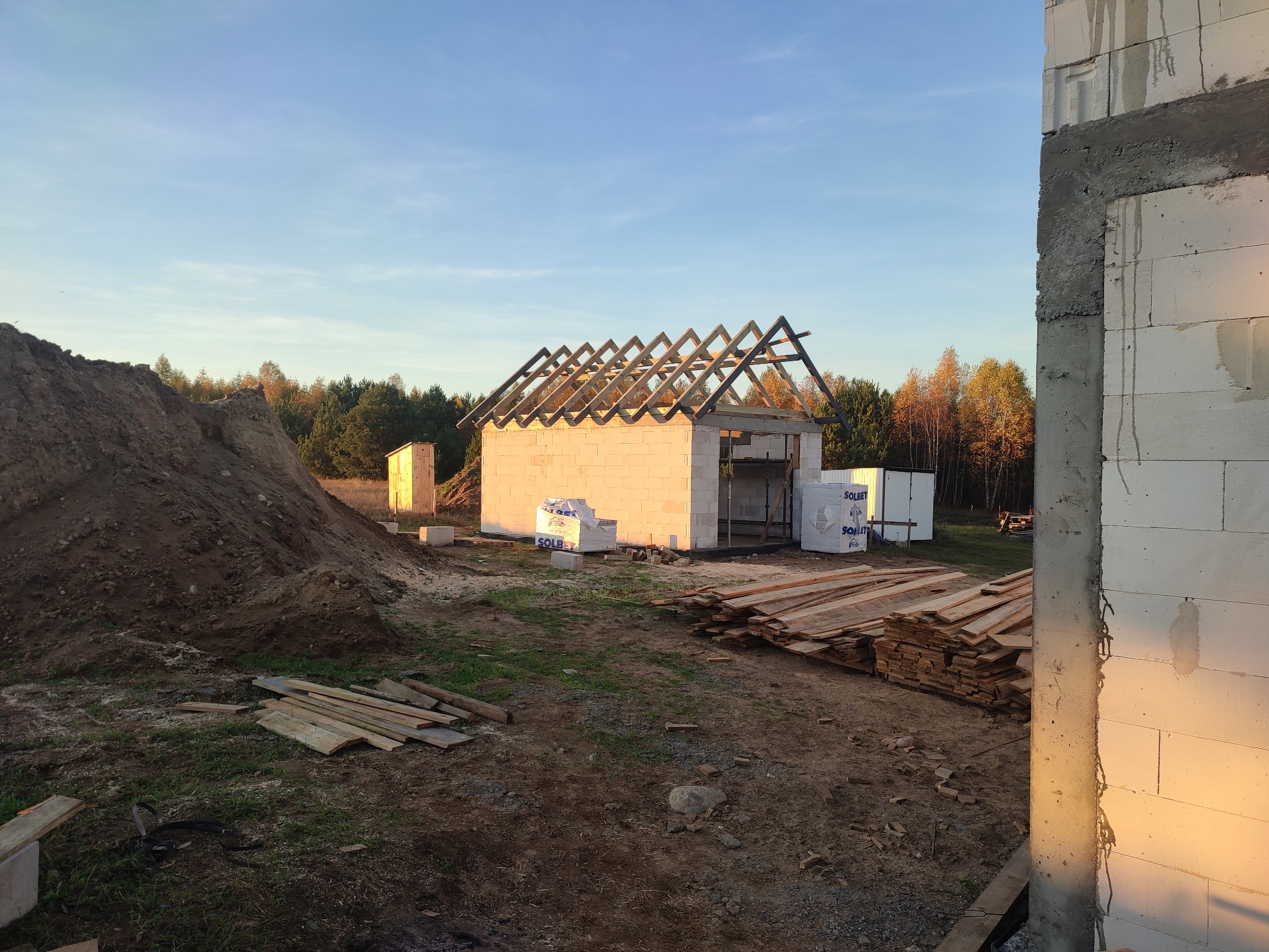 Wznoszenie parterowego budynku z bloczków betonu komórkowego; widoczne ściany, więźba dachowa, palety z bloczkami Solbet oraz sterta ziemi.
