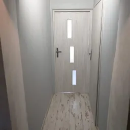 Świeżo pomalowany korytarz w odcieniach szarości z drewnopodobną podłogą i drzwiami z trzema przeszkleniami, widoczne niedoskonałości malowania przy łuku wejściowym.