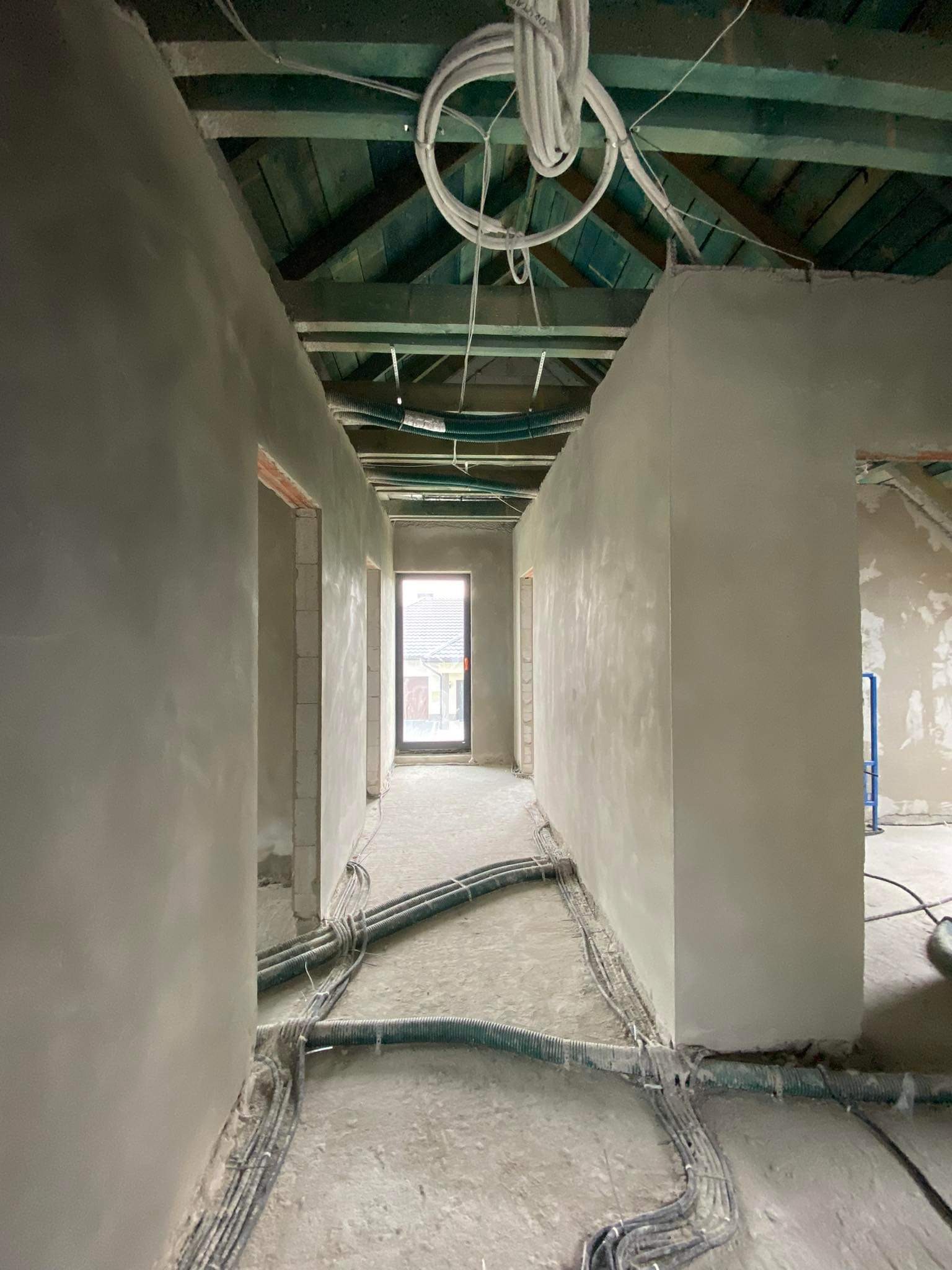 Korytarz w surowym stanie po tynkowaniu maszynowym, widoczne przewody elektryczne na podłodze i suficie, drewniany strop i otwór drzwiowy z ceglaną ramą.