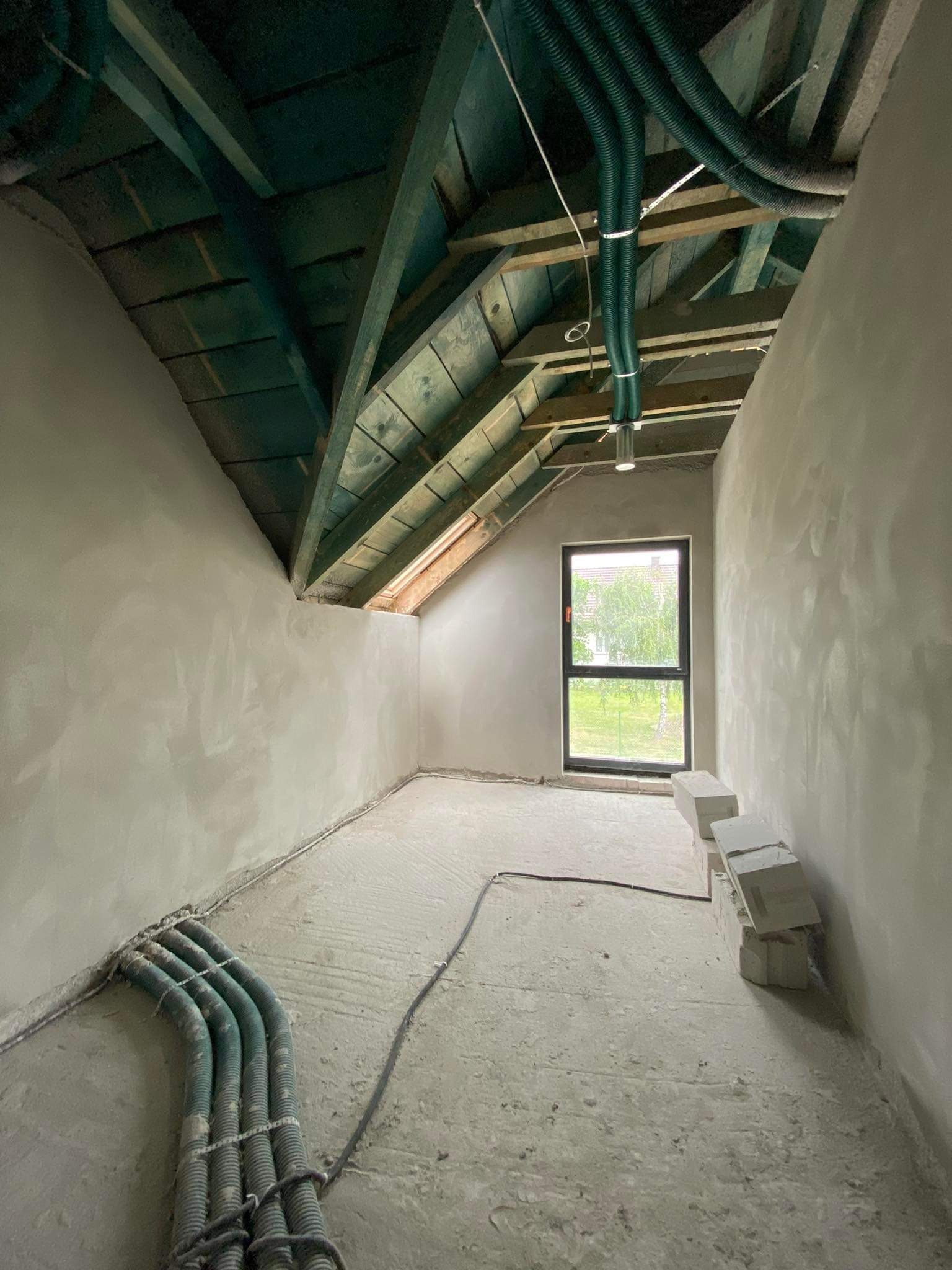 Poddasze w stanie surowym: świeżo otynkowane ściany, widoczne przewody wentylacyjne pod sufitem, okno z widokiem na zieleń i ułożone bloczki.