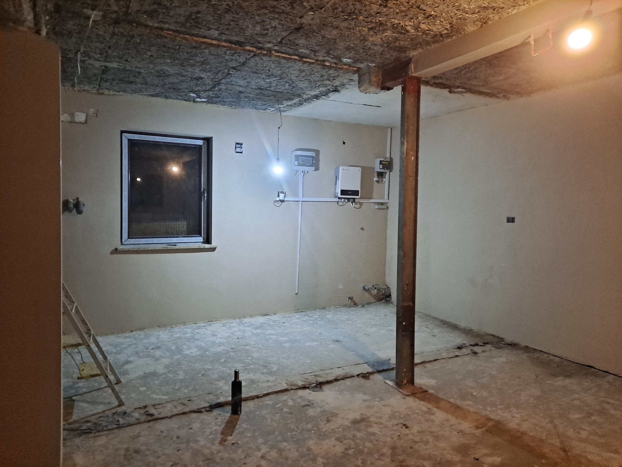 Surowe wnętrze w trakcie prac wykończeniowych: ściany po nałożeniu gładzi, widoczne instalacje elektryczne, izolacja stropu z wełny mineralnej, metalowy słup nośny, drabina oparta o ścianę, butelka...