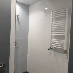 Nowoczesna łazienka z szarymi i białymi płytkami, kabina prysznicowa z panelem sterowania i biały grzejnik drabinkowy z zielonym zaworem.