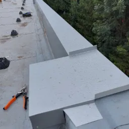 Metalowa obróbka blacharska dachu przemysłowego z widocznymi śladami deszczu, obok leżą nożyce do blachy z pomarańczowymi uchwytami.