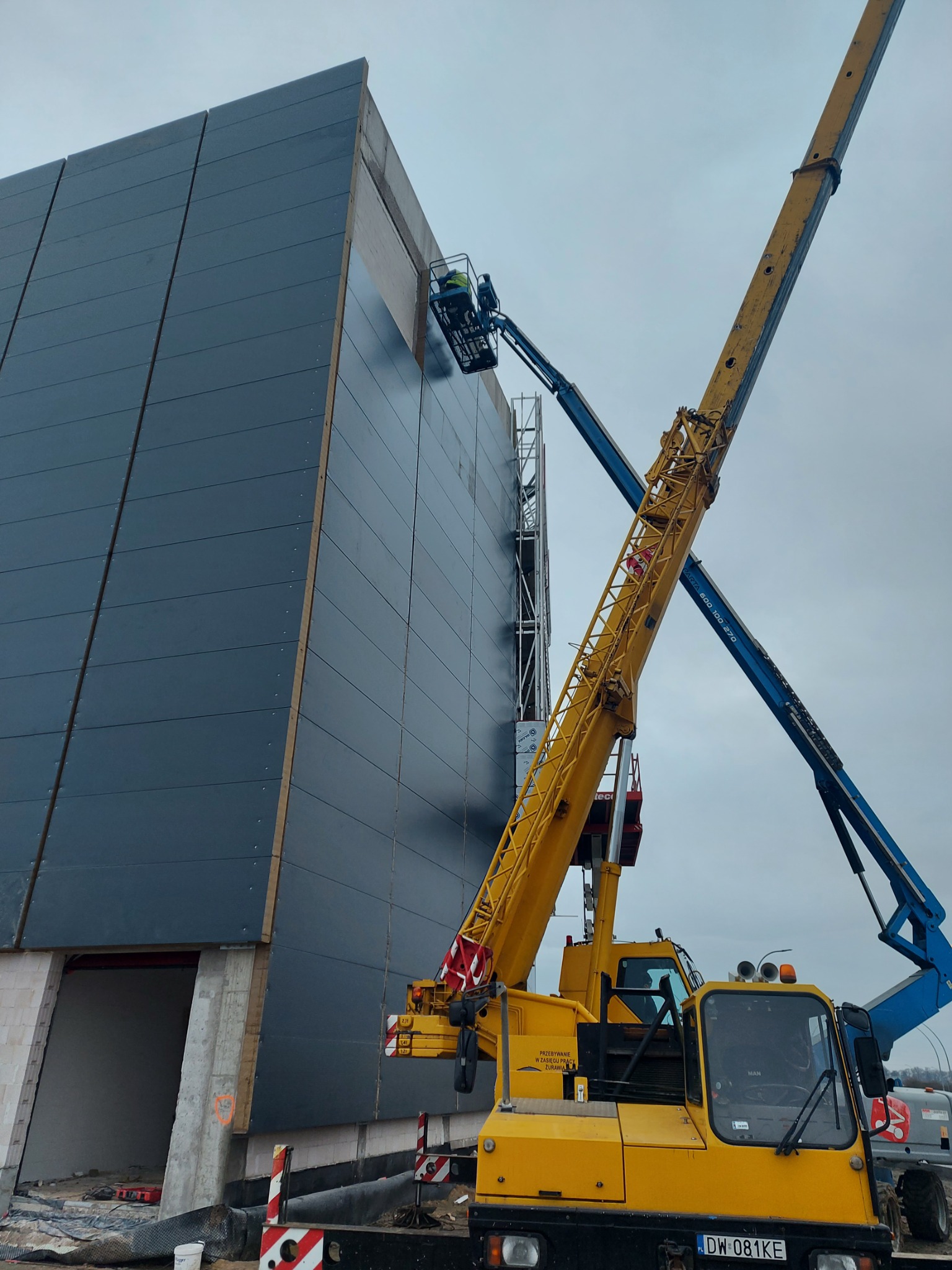 Montaż ciemnoszarych płyt warstwowych na ścianie hali przemysłowej z użyciem podnośnika koszowego i dźwigu teleskopowego w żółtym kolorze. Widoczne elementy konstrukcyjne budynku i pracownik...