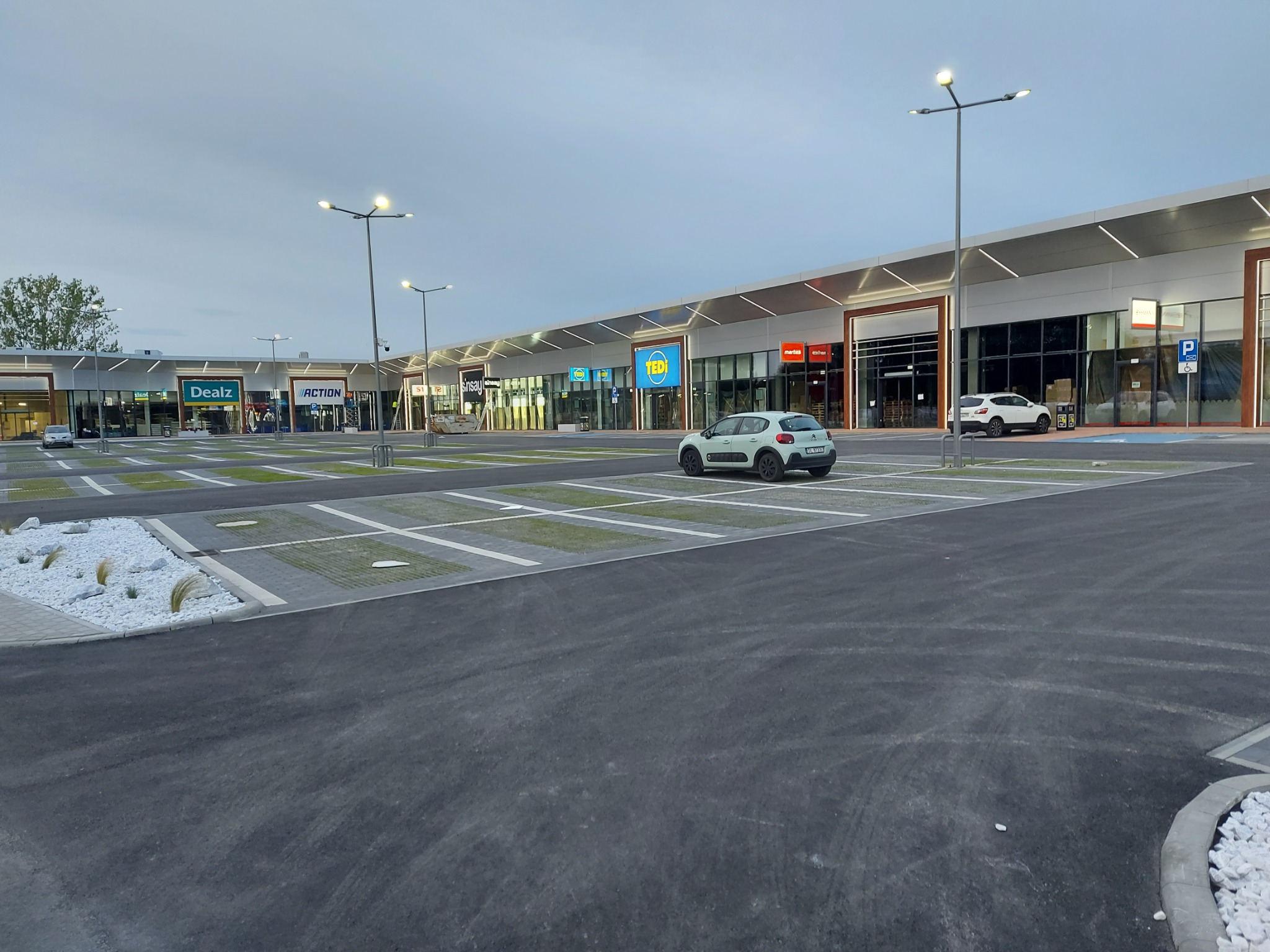 Nowoczesne centrum handlowe z parkingiem wyłożonym ażurowymi płytami i białymi kamieniami, oświetlone latarniami ulicznymi, z widocznymi szyldami sklepów.