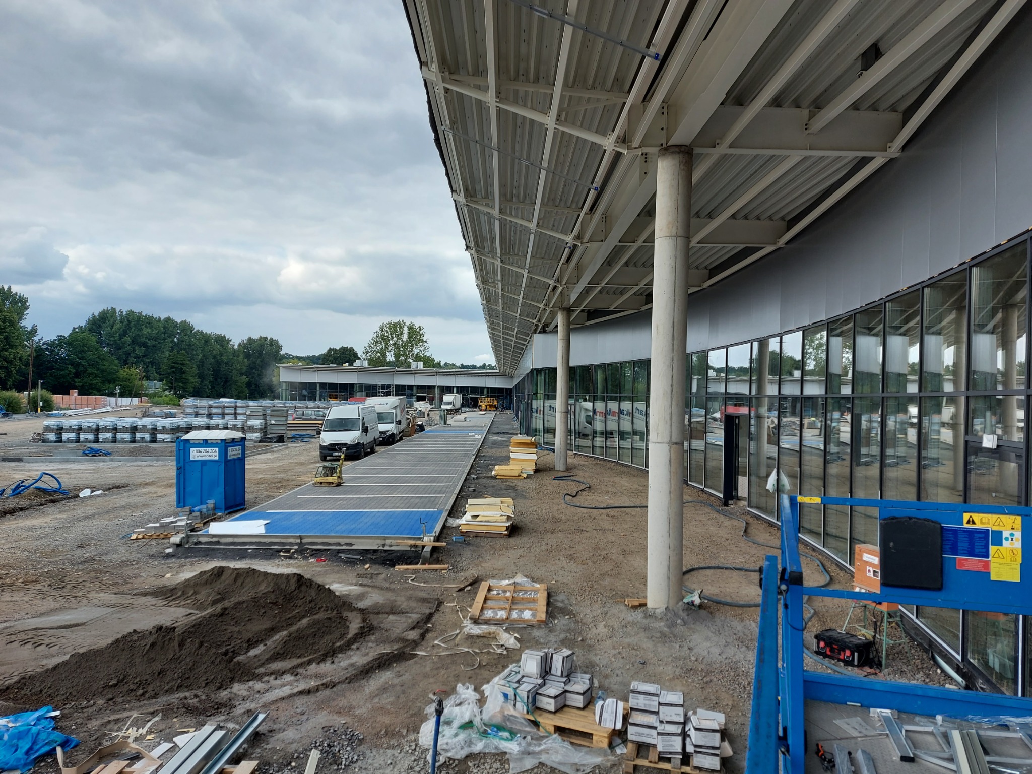 Budowa hali z płyt warstwowych w trakcie realizacji: widok na konstrukcję stalową dachu, ściany z przeszkleniami, przygotowane płyty warstwowe, zaplecze budowy z toaletą przenośną i zaparkowanymi...