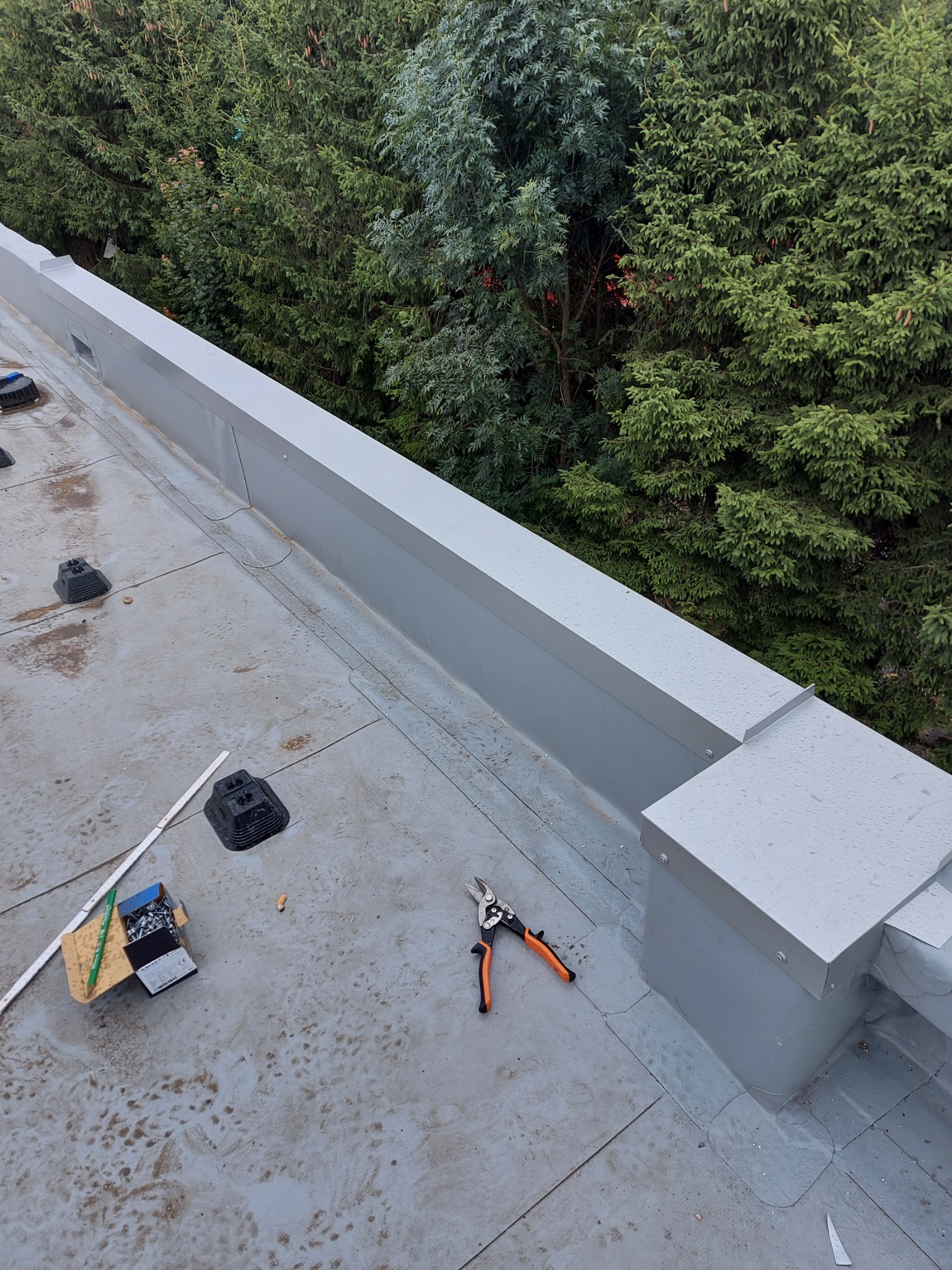 Widok z góry na fragment płaskiego dachu z zamontowaną obróbką blacharską w kolorze szarym, widoczne narzędzia dekarskie, elementy mocujące oraz podkładki pod instalacje na tle zieleni drzew.