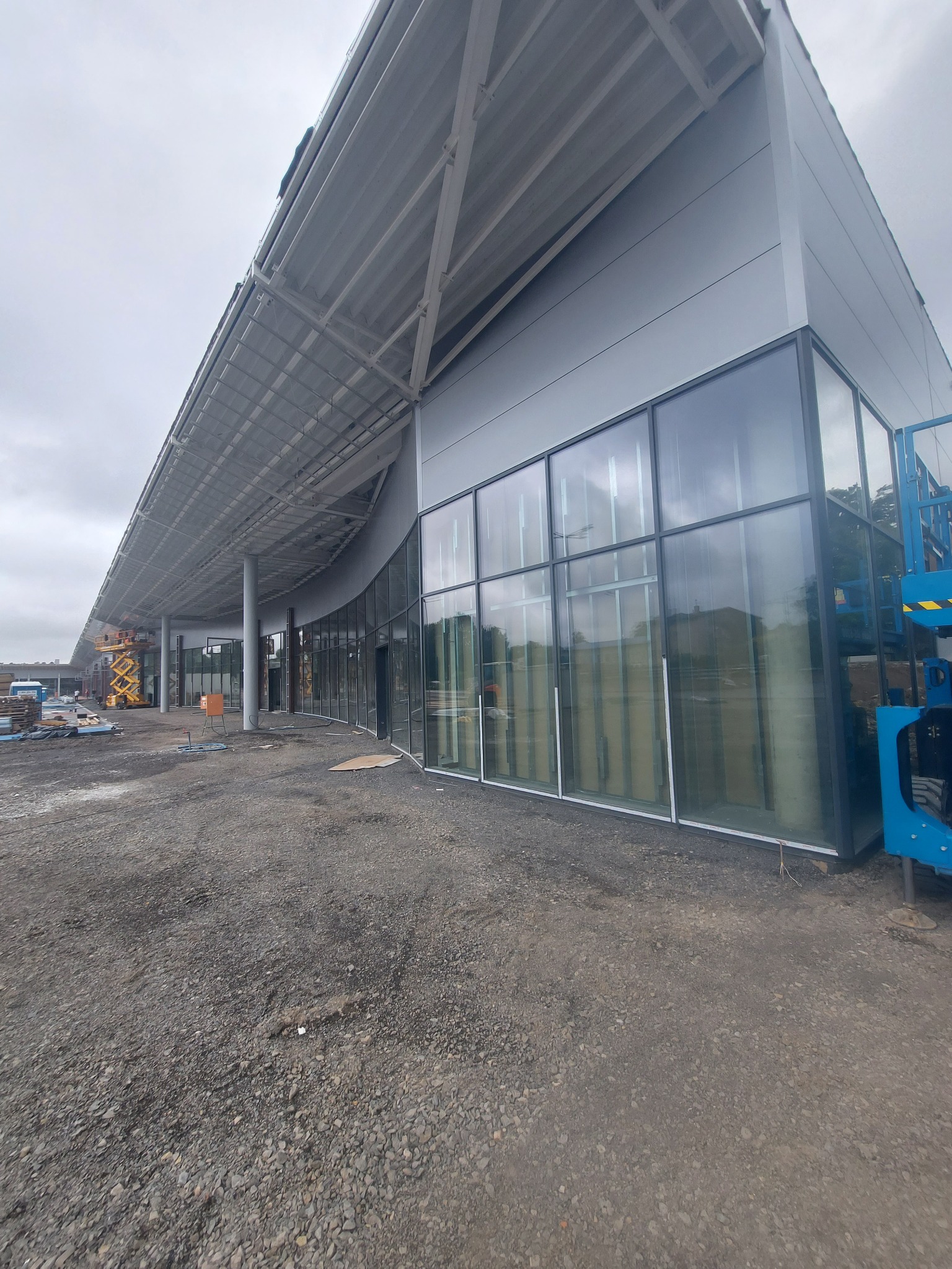 Budowa hali: Widok z zewnątrz na konstrukcję hali z dużymi oknami, widoczny szary panelowy siding, stalowa konstrukcja dachu, teren budowy i podnośnik koszowy w tle.