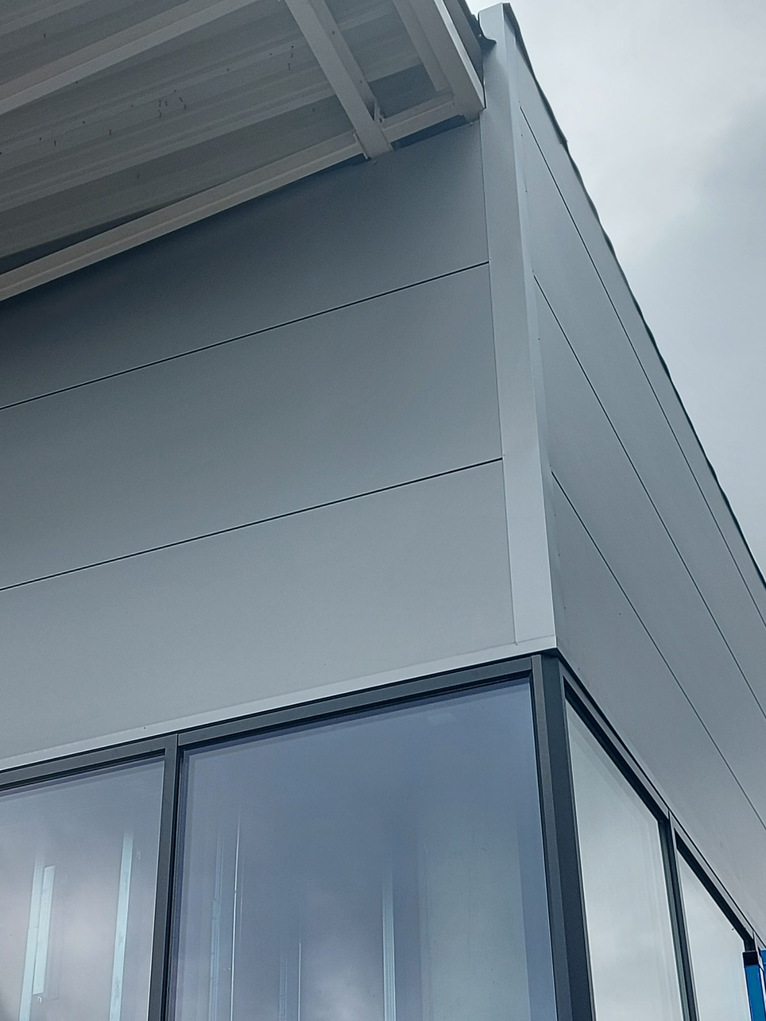 Fragment narożnika budynku przemysłowego z szarymi płytami warstwowymi i dużymi oknami z czarnymi ramami, widok z dołu na tle pochmurnego nieba.