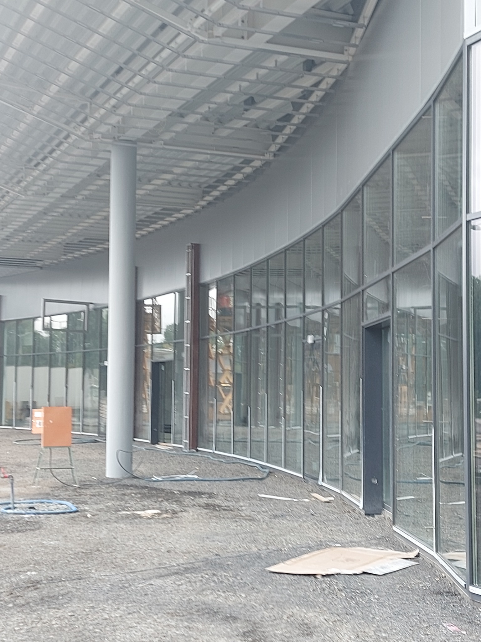 Budowa hali: widok na zakrzywioną fasadę z paneli szklanych i stalową konstrukcję dachu, z widocznymi elementami rusztowania w tle i tymczasowymi akcesoriami na placu budowy.