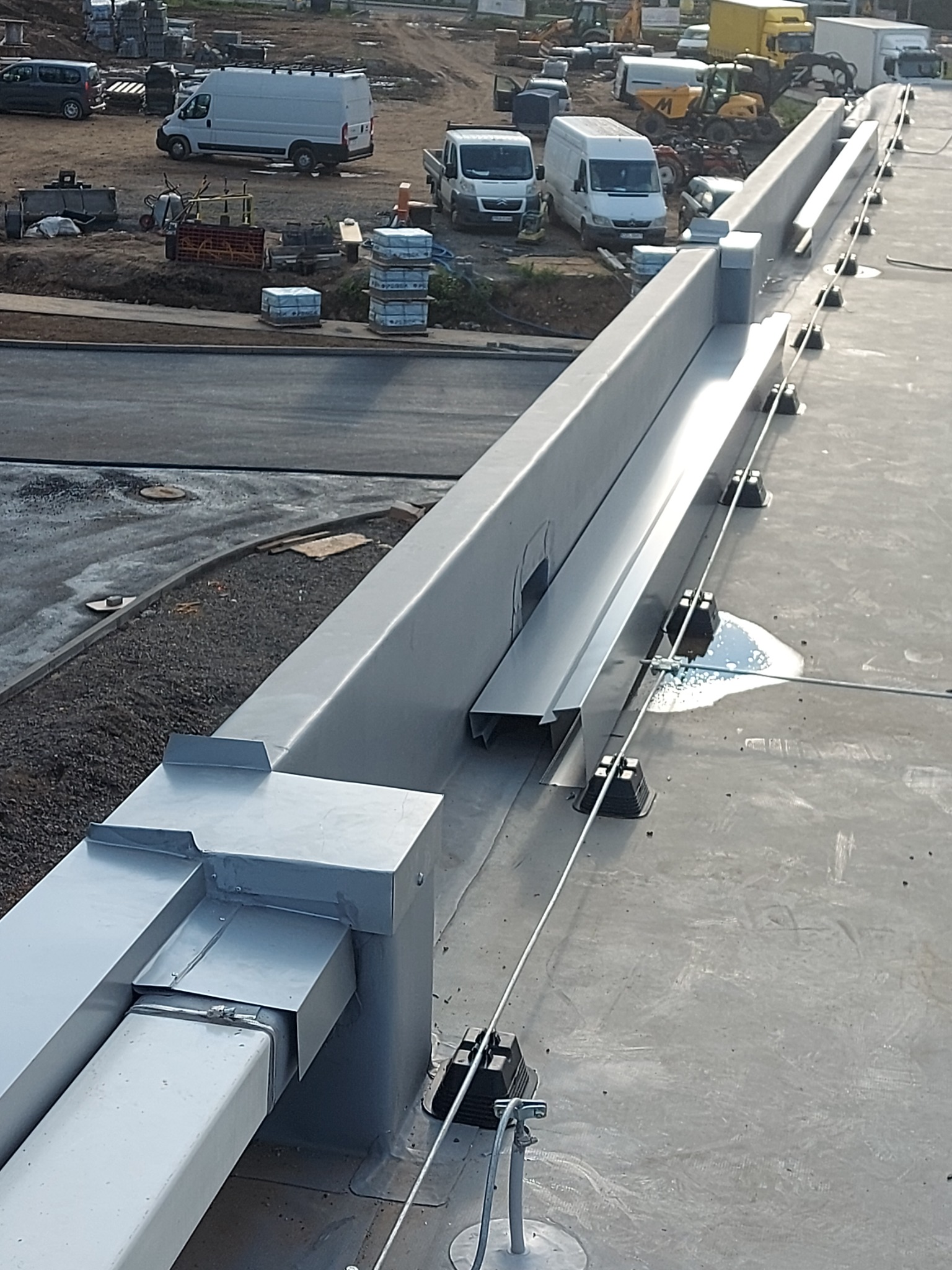 Szare, metalowe rynny dachowe na płaskim dachu hali przemysłowej z widocznymi elementami mocującymi i linkami zabezpieczającymi, w tle teren budowy z maszynami i pojazdami.