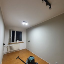 Kompleksowe wykończenia wnętrz Łukasz Sawicki - Pokój po malowaniu z drewnianą podłogą, widoczny odkurzacz przemysłowy i grzejnik pod oknem z widokiem na nocne miasto.