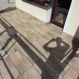 Kompleksowe wykończenia wnętrz Łukasz Sawicki - Wyłożony płytkami balkon z drewnopodobnym wzorem, widoczny cień osoby robiącej zdjęcie, brązowa balustrada i fragment ściany z oknem, na parapecie stoi kubek.