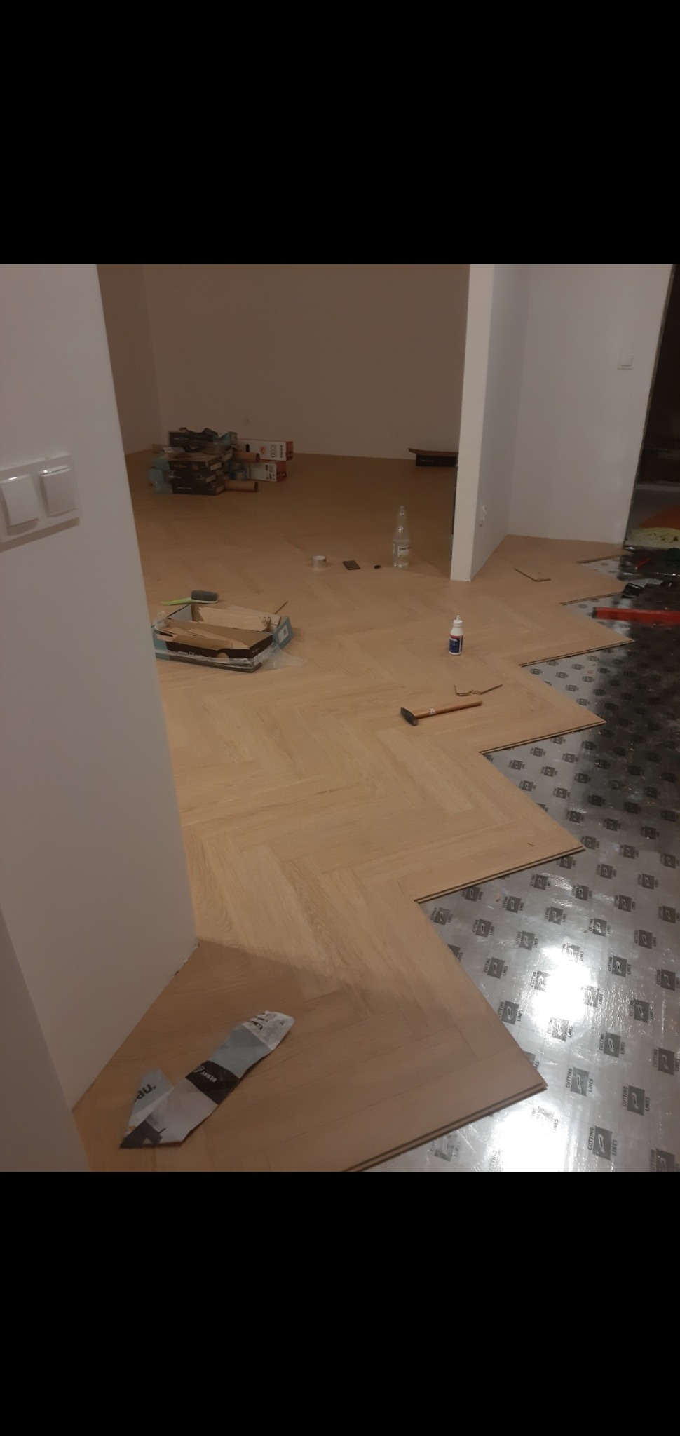 Układanie parkietu w jodełkę w trakcie prac. Część podłogi wyłożona folią izolacyjną, widoczne narzędzia i klej.